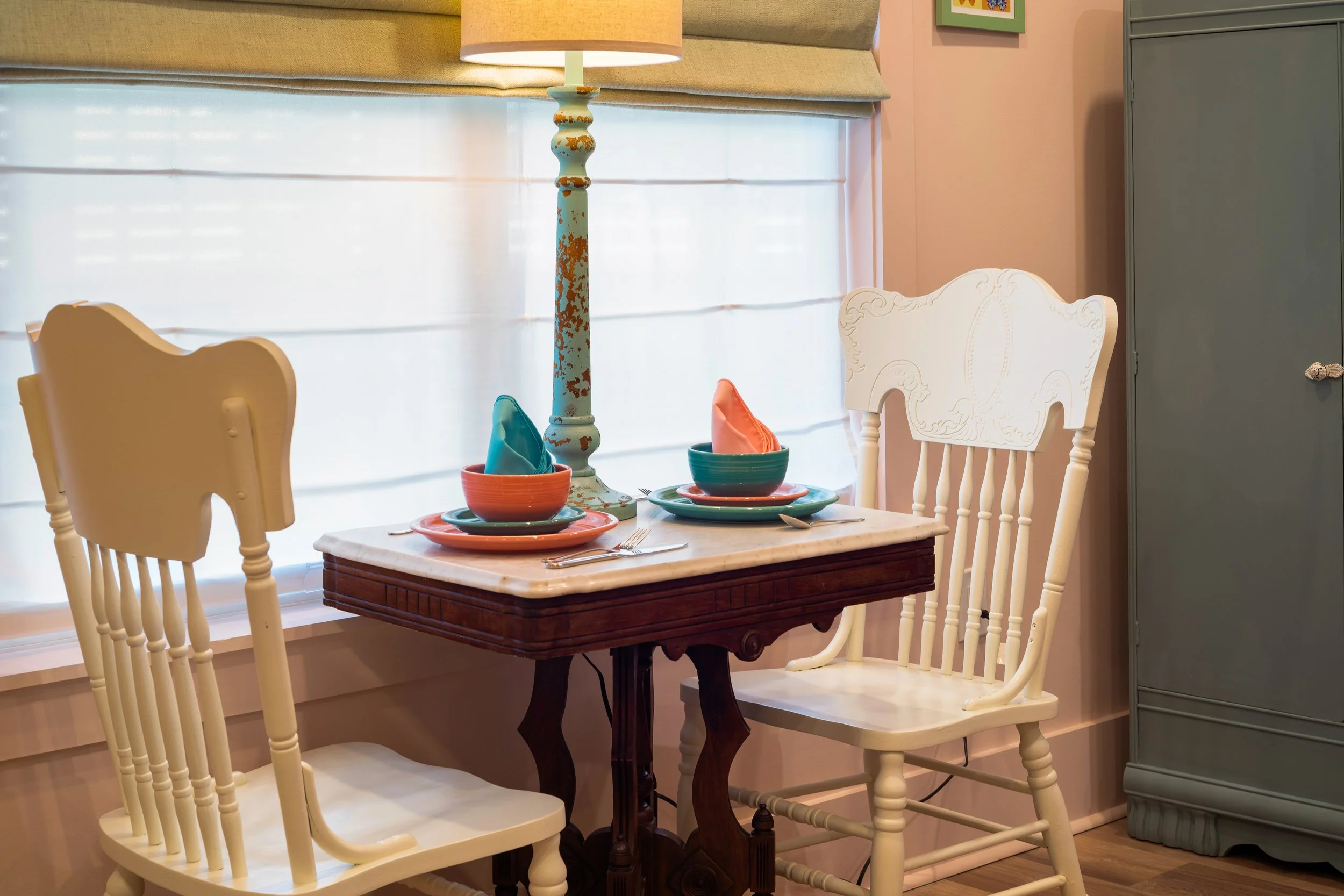 A dining table set with colorful dishes and napkins, surrounded by white wooden chairs, next to a window with blinds and a vintage lamp on the table.