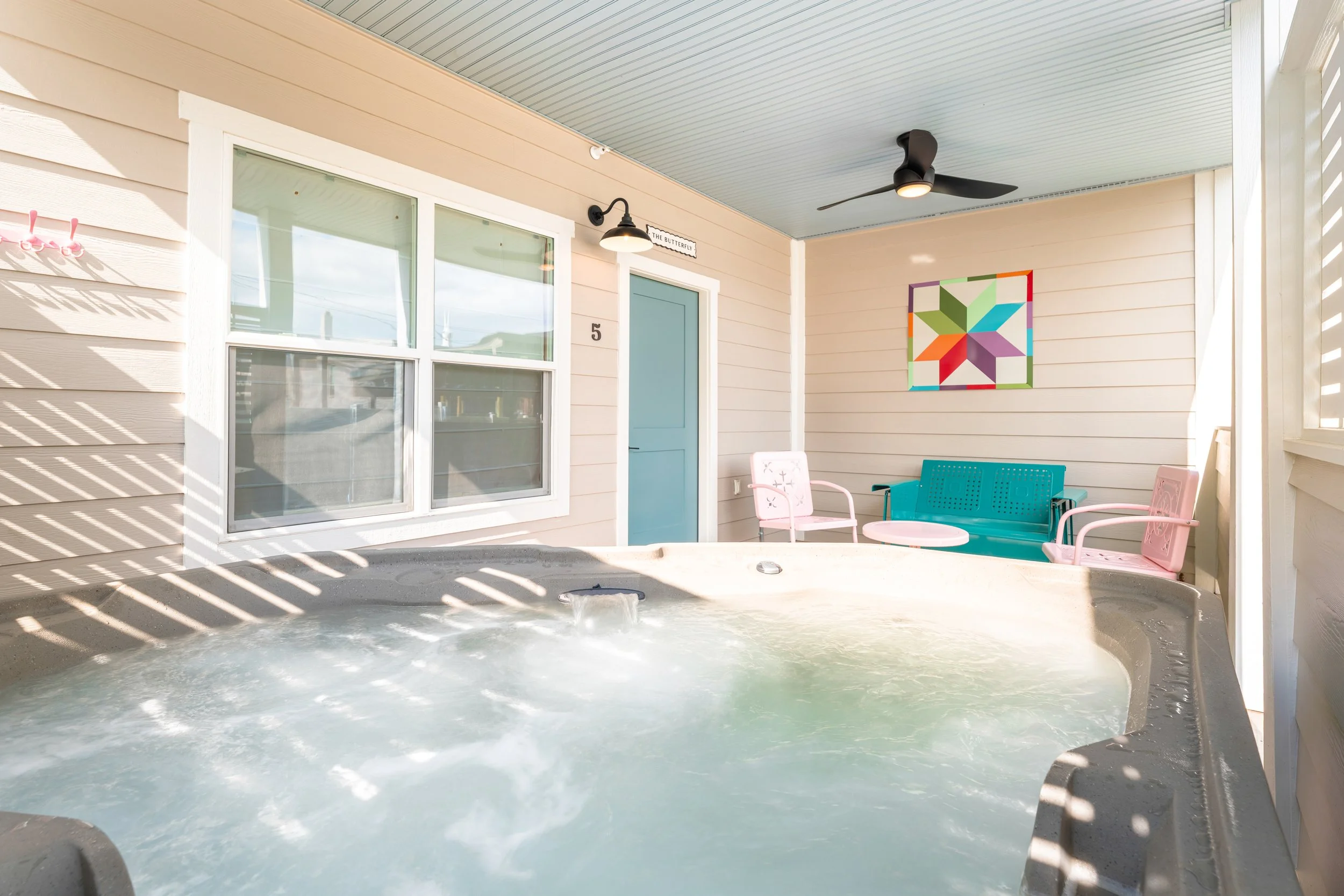 A covered outdoor patio with a small pool or hot tub filled with water and bubbles. There is a blue door, pink chairs, a teal bench, a white table, colorful wall art, outdoor wall lights, and a ceiling fan.