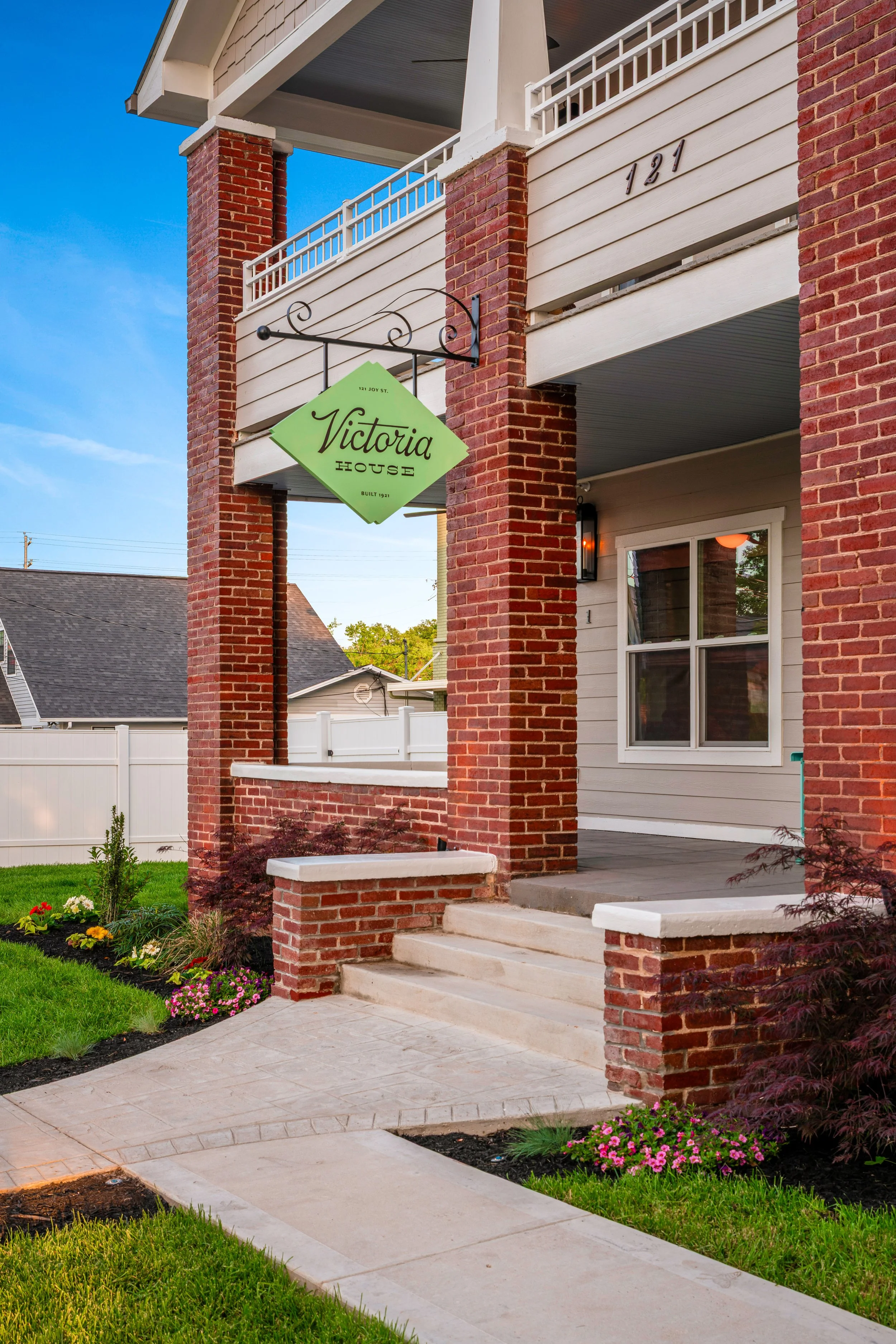 The front porch of a house with red brick columns and beige siding, featuring a hanging green sign that says 'Victoria House' and a well-maintained garden with colorful flowers and green grass.