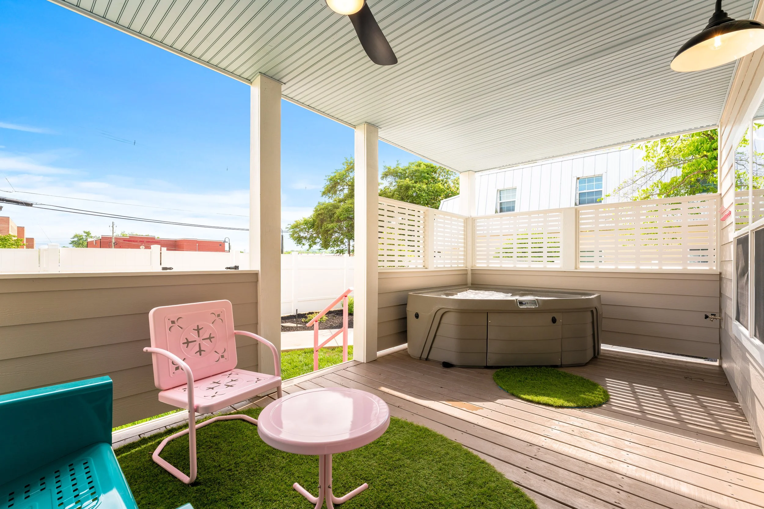 Covered patio with a hot tub, pink and blue chairs, and outdoor furniture, overlooking a neighborhood with trees and blue sky.