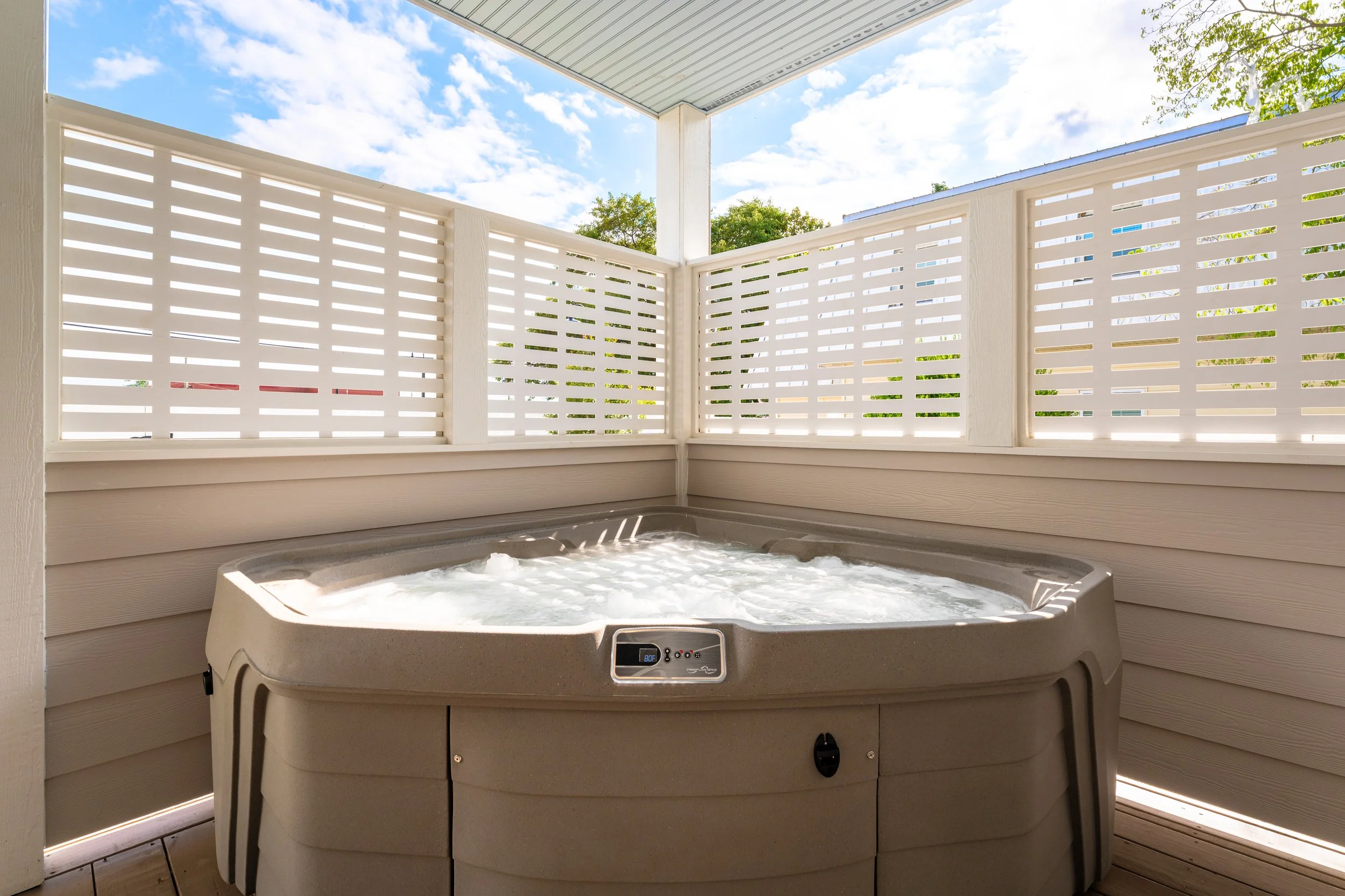 A hot tub on a porch, with white lattice fencing and a view of the blue sky and trees outside.