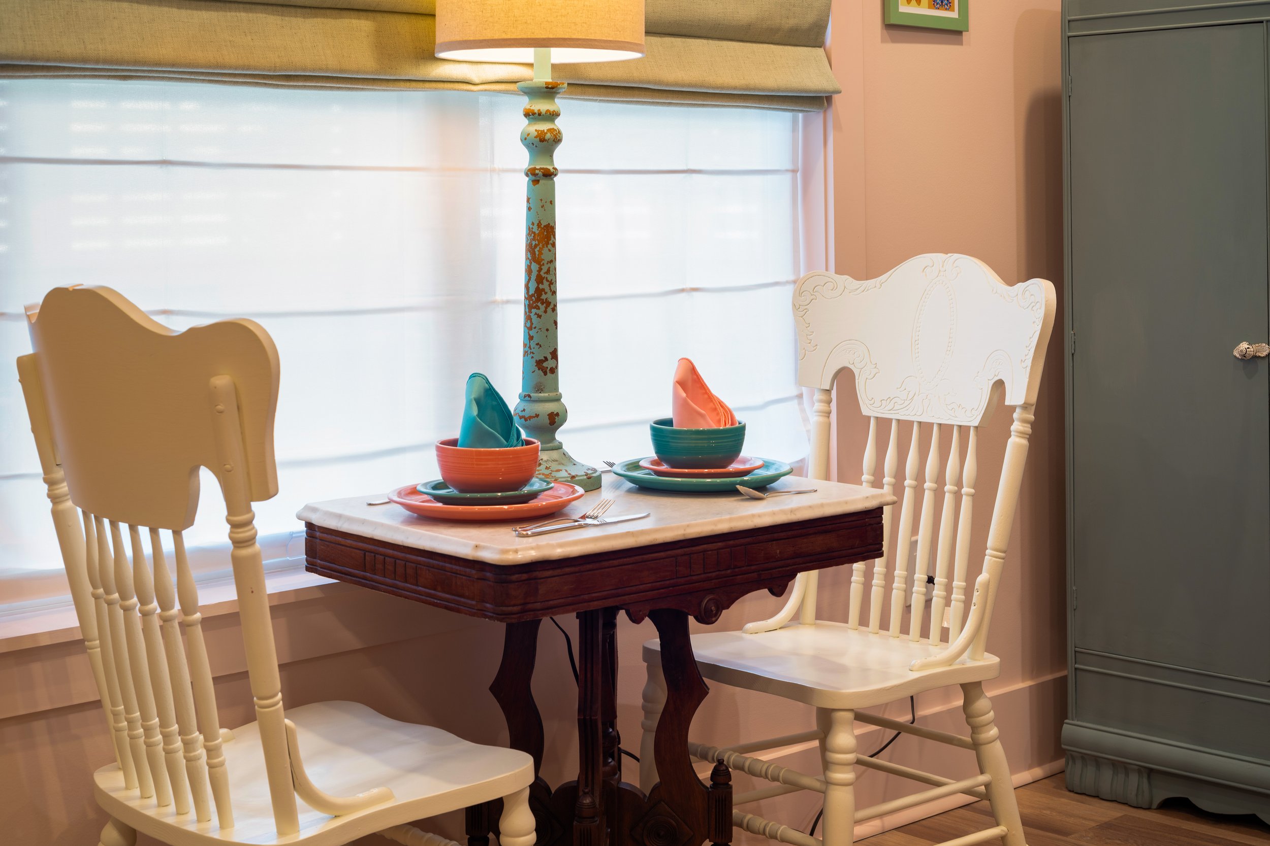 A dining room table set with colorful bowls, plates, and napkins, with a vintage-style lamp and chairs around the table. There's a window with blinds and a gray cabinet in the background.