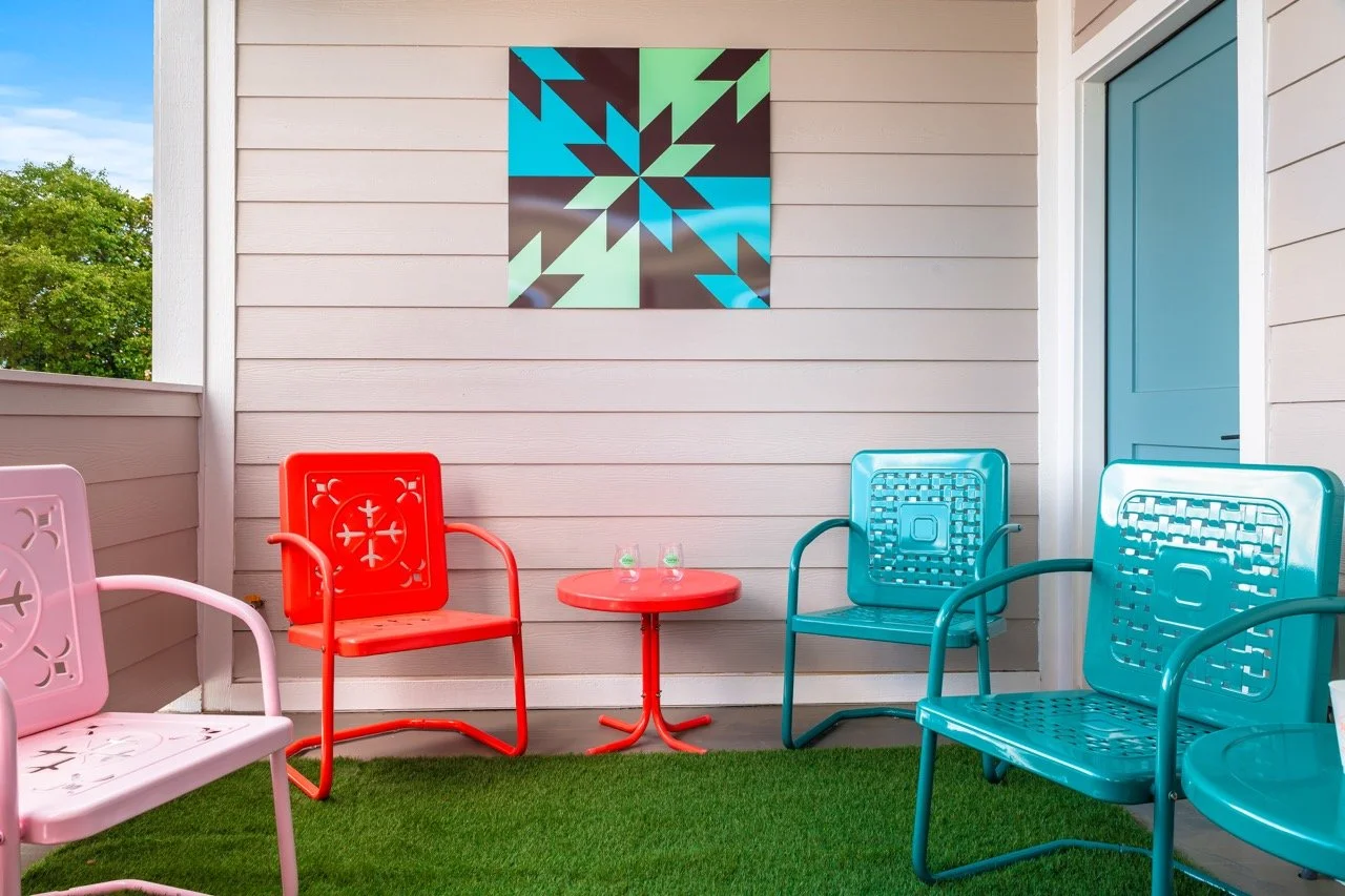 Balcony with colorful metal chairs in pink, red, and blue, a red round table with two glasses, white horizontal siding wall, and a modern geometric art piece on the wall.