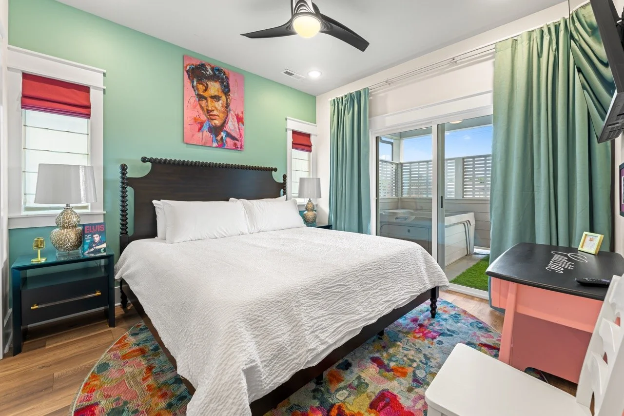 A bedroom with a large bed, white bedding, and a black wooden headboard. There are two windows with white frames, red Roman shades, and white blinds. A nightstand with a white lamp, a small picture, and a decorative item is on each side of the bed. A