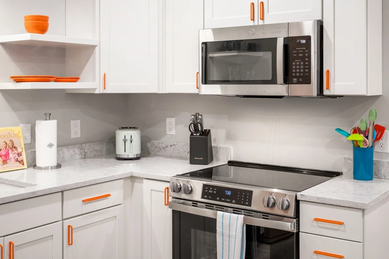 Modern kitchen with white cabinets, stainless steel oven and microwave, marble countertops, and colorful utensils in a blue holder.