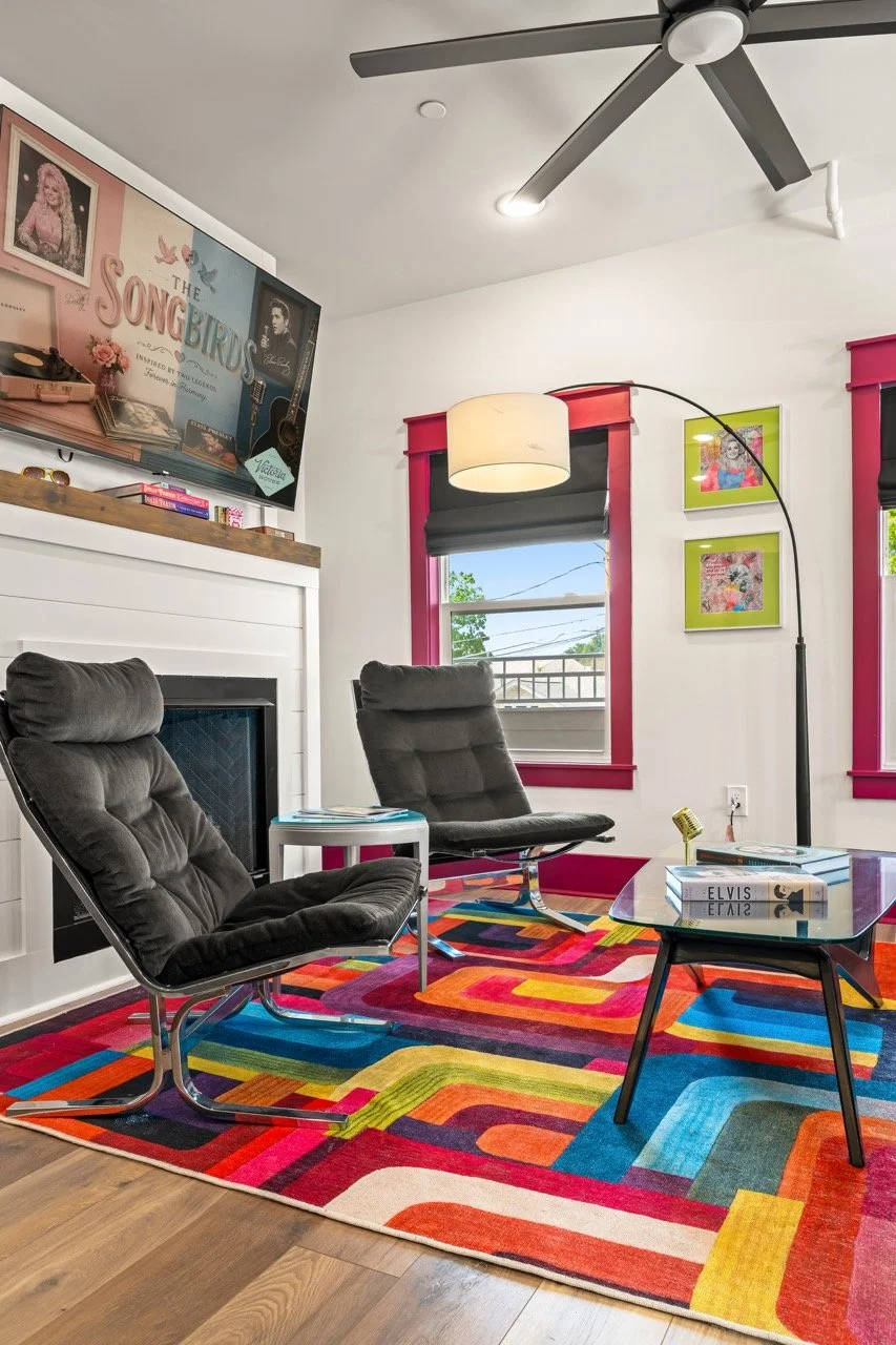 Living room with two black chairs, a colorful geometric rug, a white side table with books, a modern arc floor lamp, a window with pink and gray trim, and a colorful wall art display featuring a record, a guitar, and framed pictures.