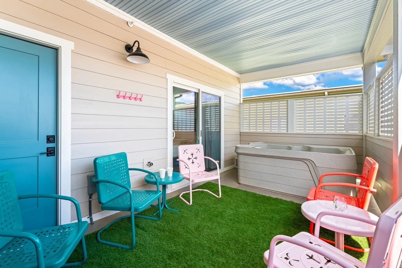 Covered patio with colorful chairs, a hot tub, and a small table with cups, on artificial grass with a beige exterior wall and a sliding glass door.