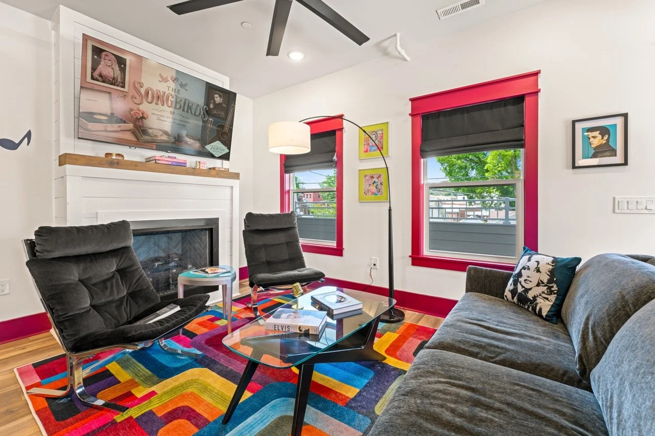 Living room with white walls, two black armchairs, a gray sofa with a Marilyn Monroe pillow, a colorful geometric rug, a glass coffee table with books, trailing lamp, fireplace with a wooden mantel, two large windows with pink borders and black blind