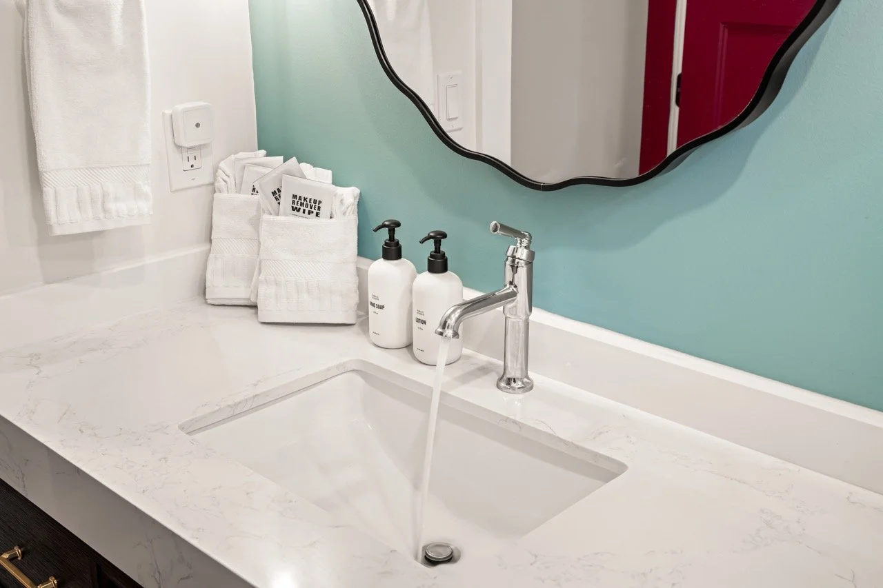 Bathroom sink with water running from the faucet, soap and lotion bottles, neatly folded white towels, a mirror, and a wall outlet on a white marble countertop.