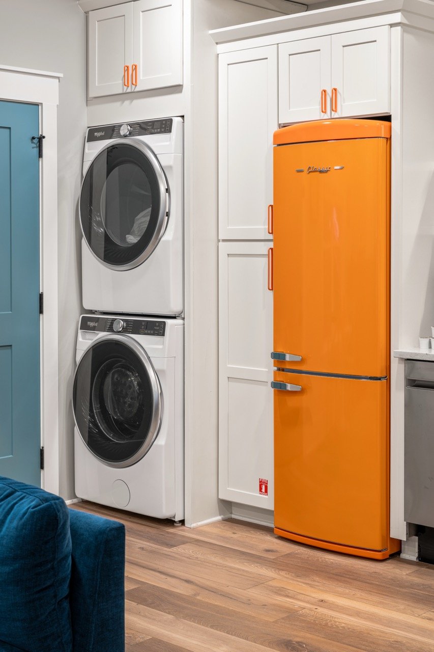 A laundry area with stacked white washer and dryer, white cabinets above, and an orange retro-style refrigerator on the right. Part of a blue door and wooden floor are also visible.