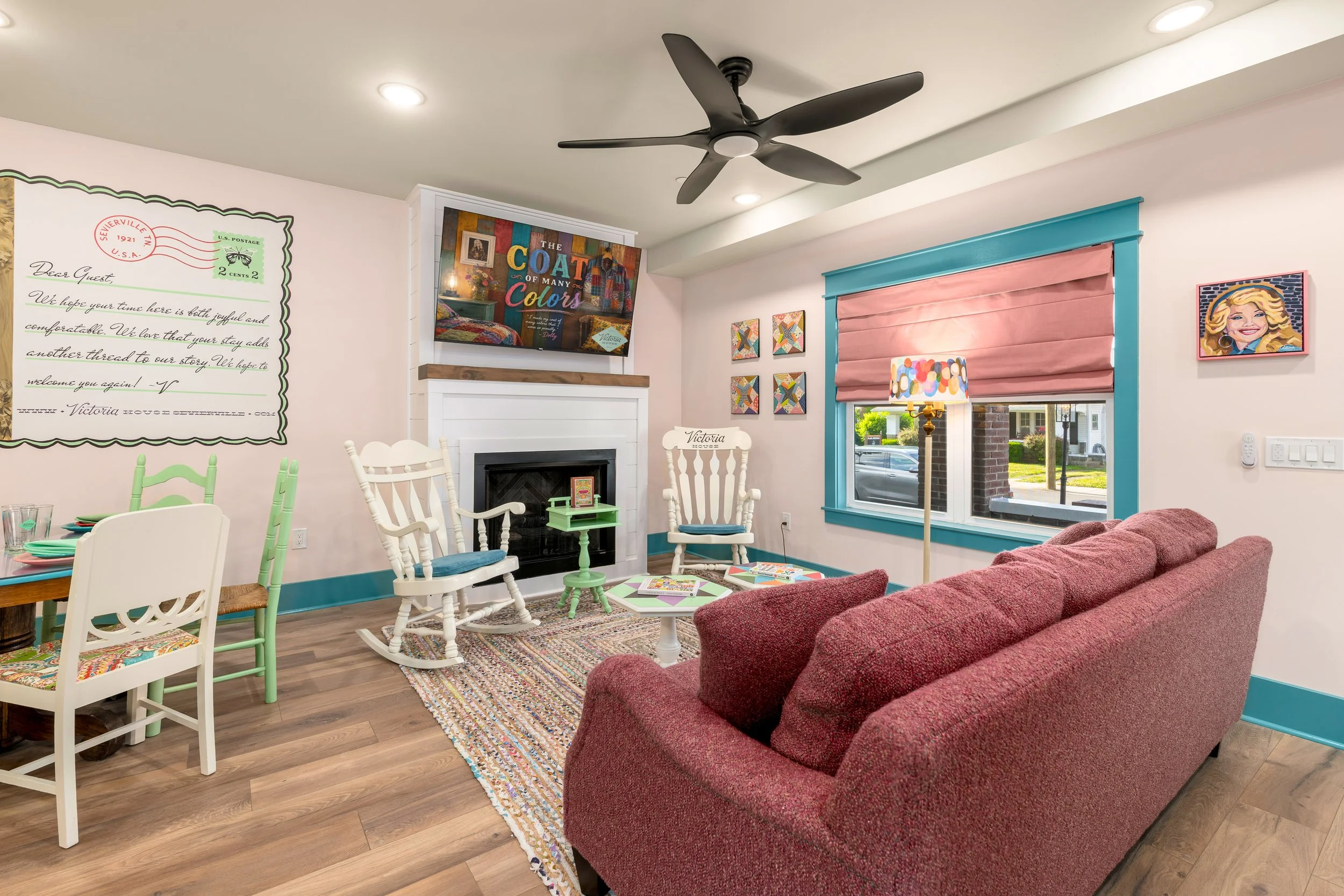 A cozy living room with a red upholstered sofa, white and pastel-colored chairs, a colorful area rug, a fireplace, framed artwork, a large window with pink shades, and a ceiling fan.