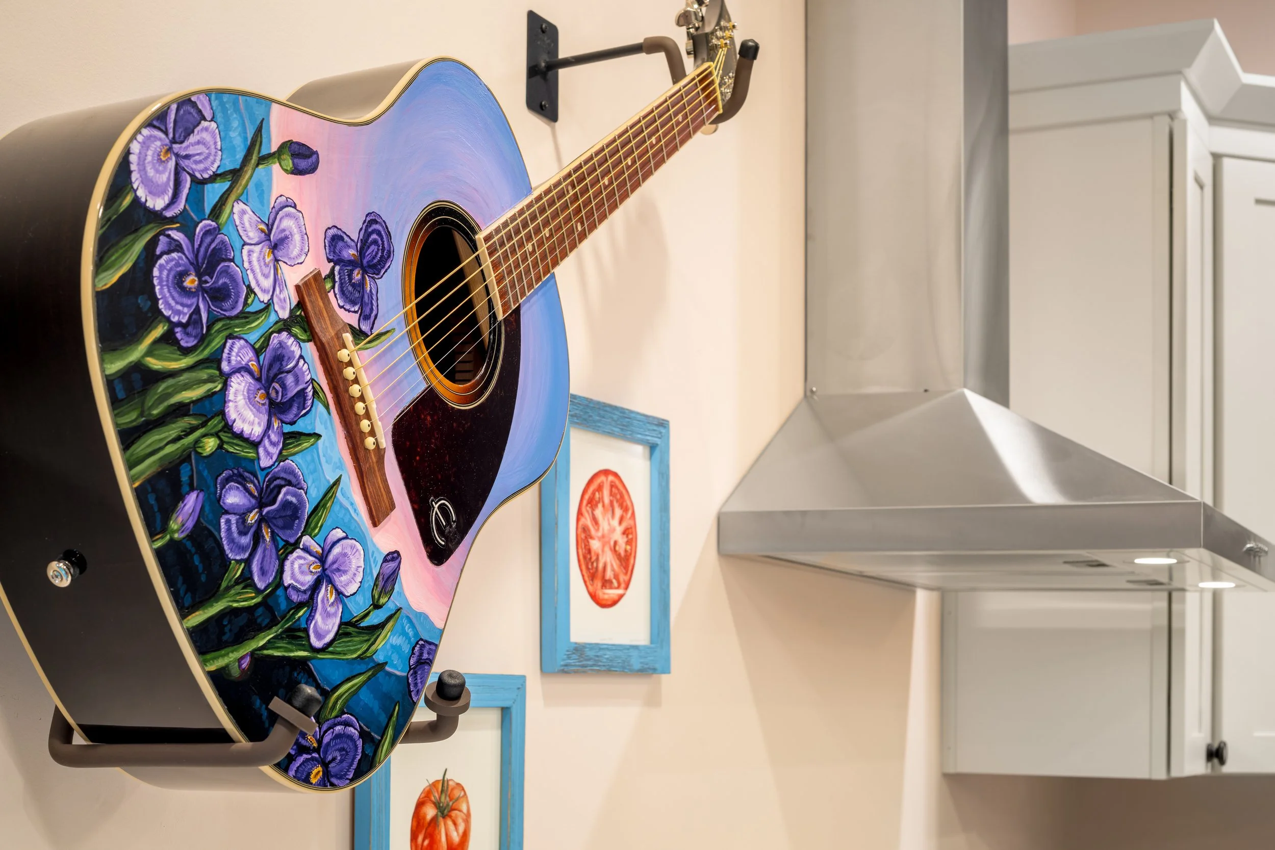 A colorful acoustic guitar with purple iris flower artwork on the body, sitting on a black guitar stand in a room with a white wall, a white cabinet, and a metal vent hood.