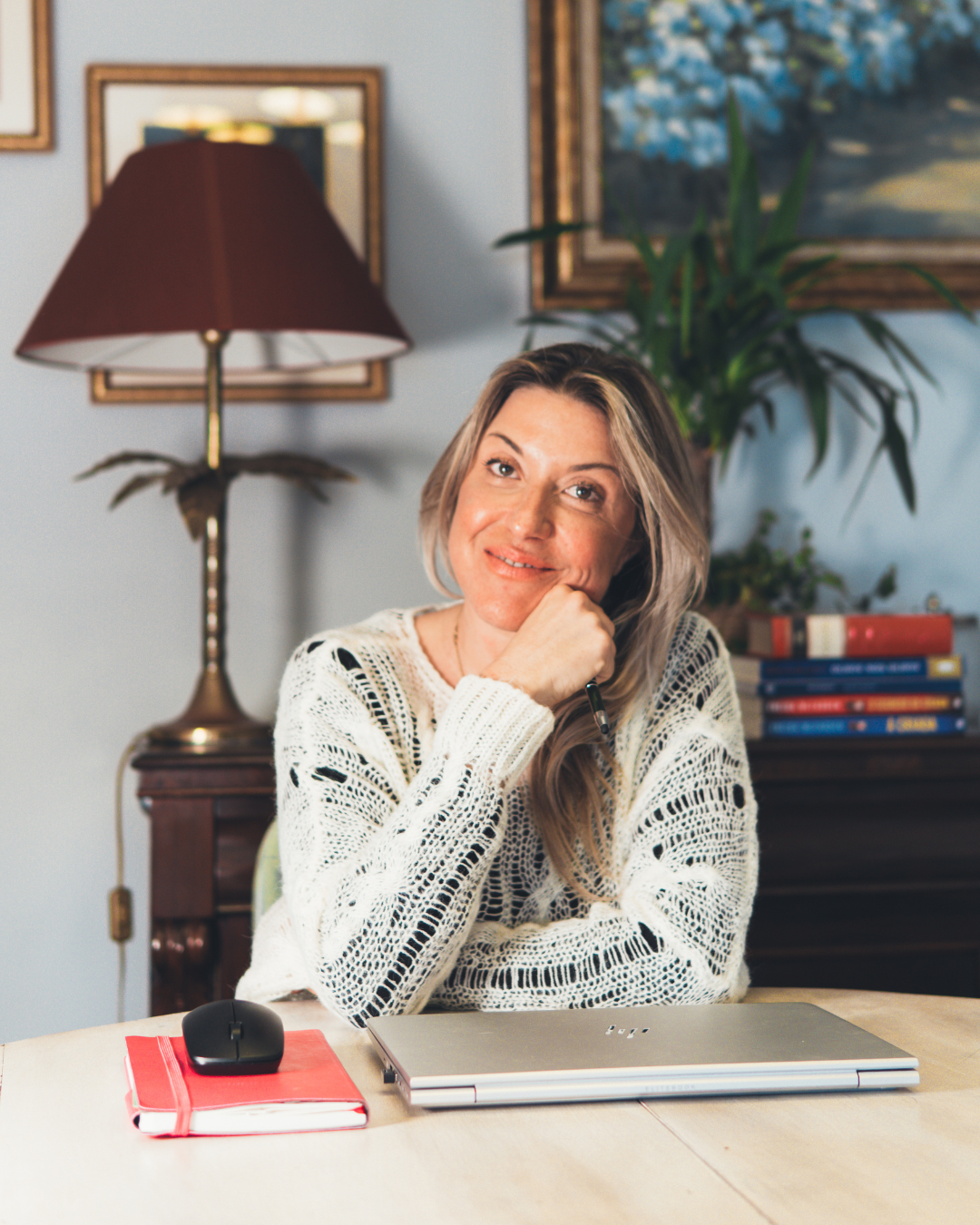 Mulher com cabelo loiro, usando blusa de tricô branca, sentada em uma mesa com um laptop, um mouse, um caderno vermelho e um cachorro pensativo, em um ambiente com uma luminária de aba marrom, quadros na parede e uma planta de folhas verdes ao fundo.