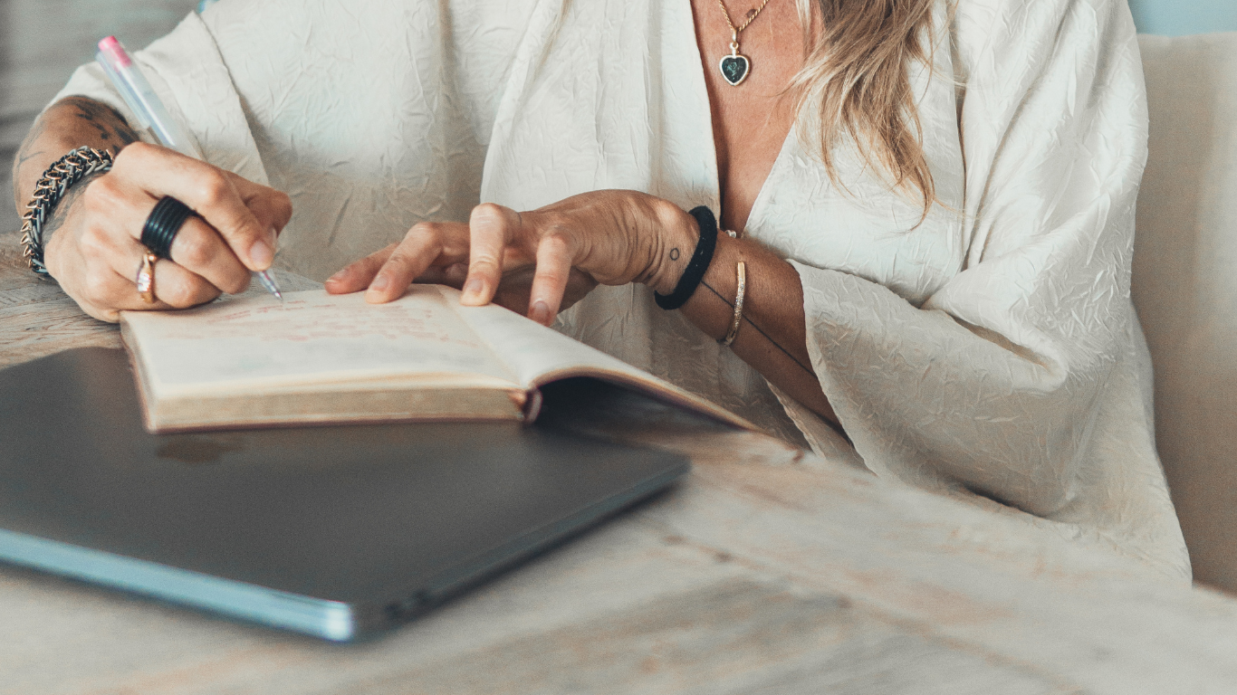 Pessoa escrevendo em um caderno, com um laptop na mesa, usando roupas brancas, acessórios e joias.