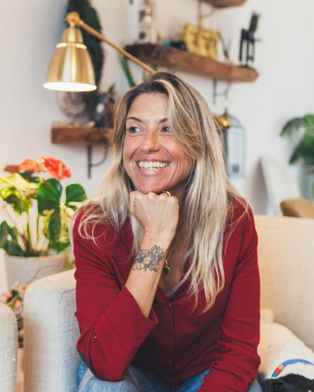 Mulher sorridente com cabelo loiro, camisa vermelha, tatuagem no braço, sentada em um sofá, em ambiente decorado com plantas, prateleiras de madeira e iluminação moderna.