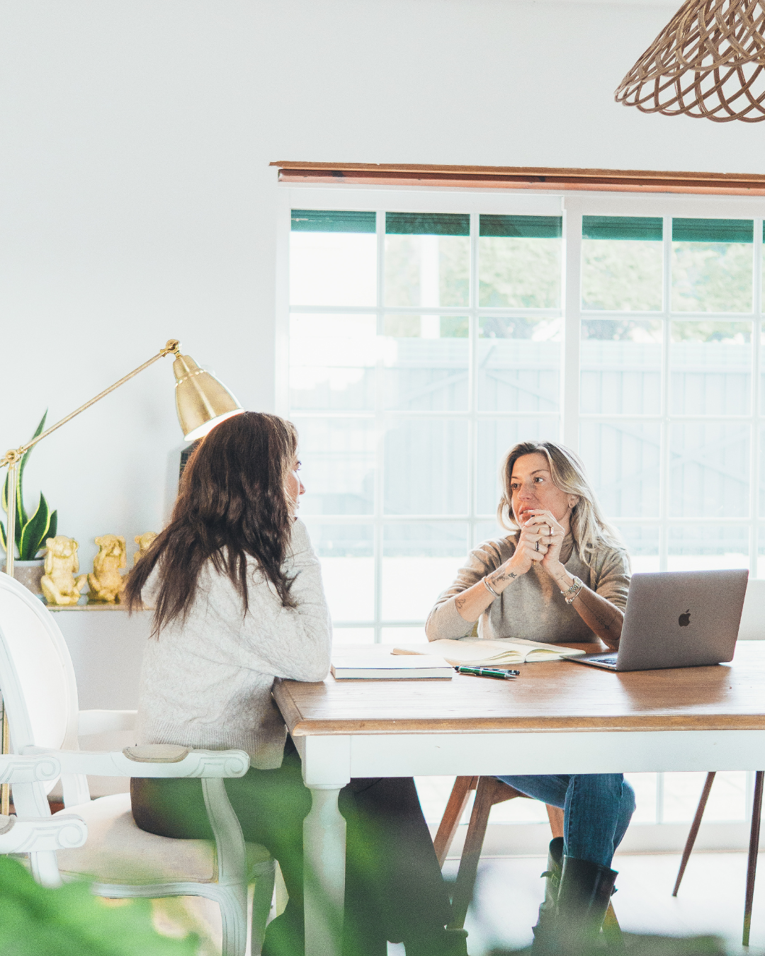 Duas mulheres conversando em uma mesa de escritório, com computador portátil, caderno e canetas, em ambiente bem iluminado com janela grande ao fundo.
