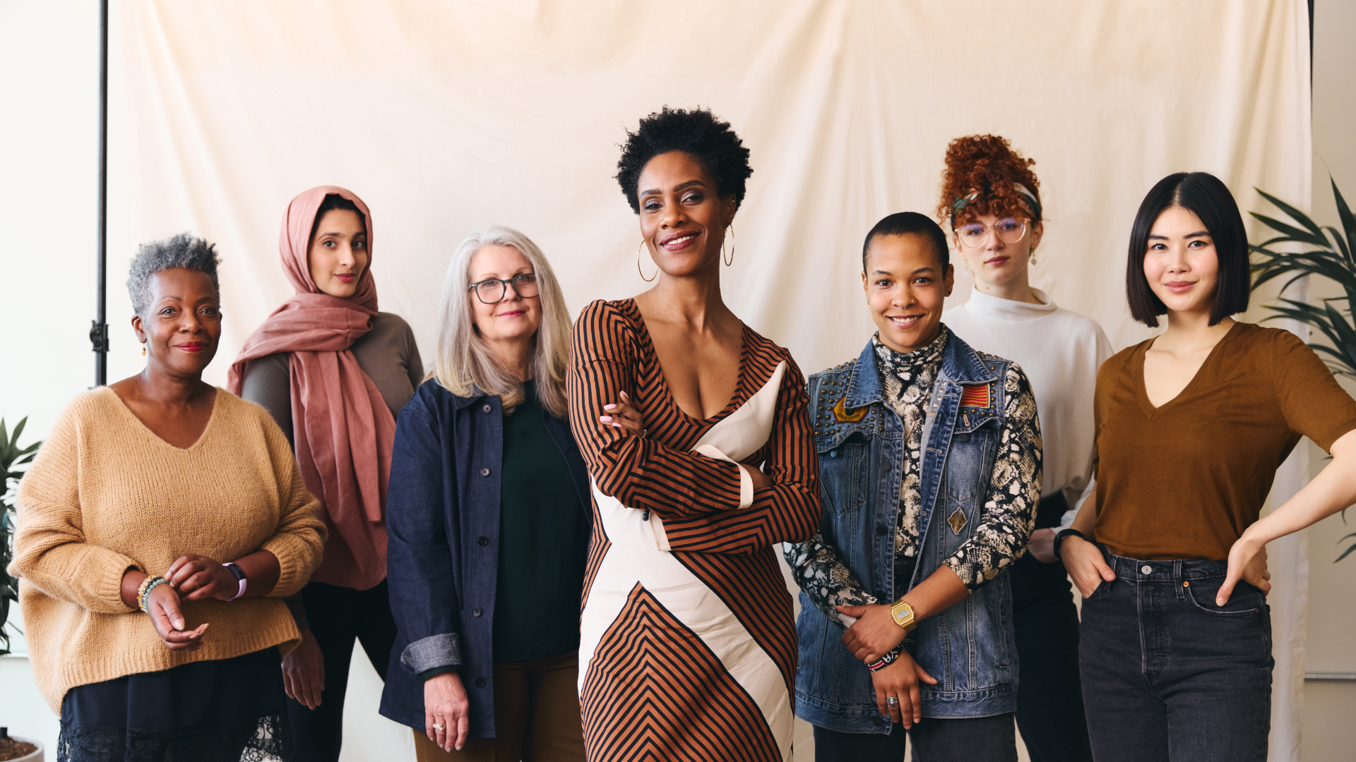 A diverse group of seven women standing together indoors in front of a beige backdrop, smiling and posing with confidence for the photo.
