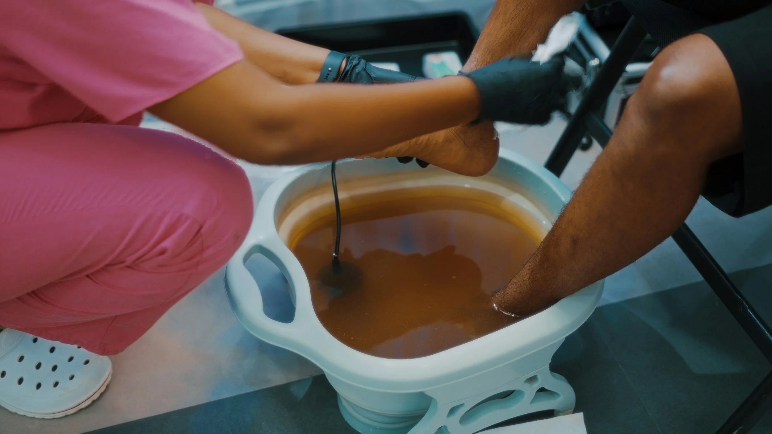 Two people are using a foot spa machine, with their legs submerged in water, during a pedicure treatment. One person is wearing pink pants and white sneakers, while the other person's legs are bare.