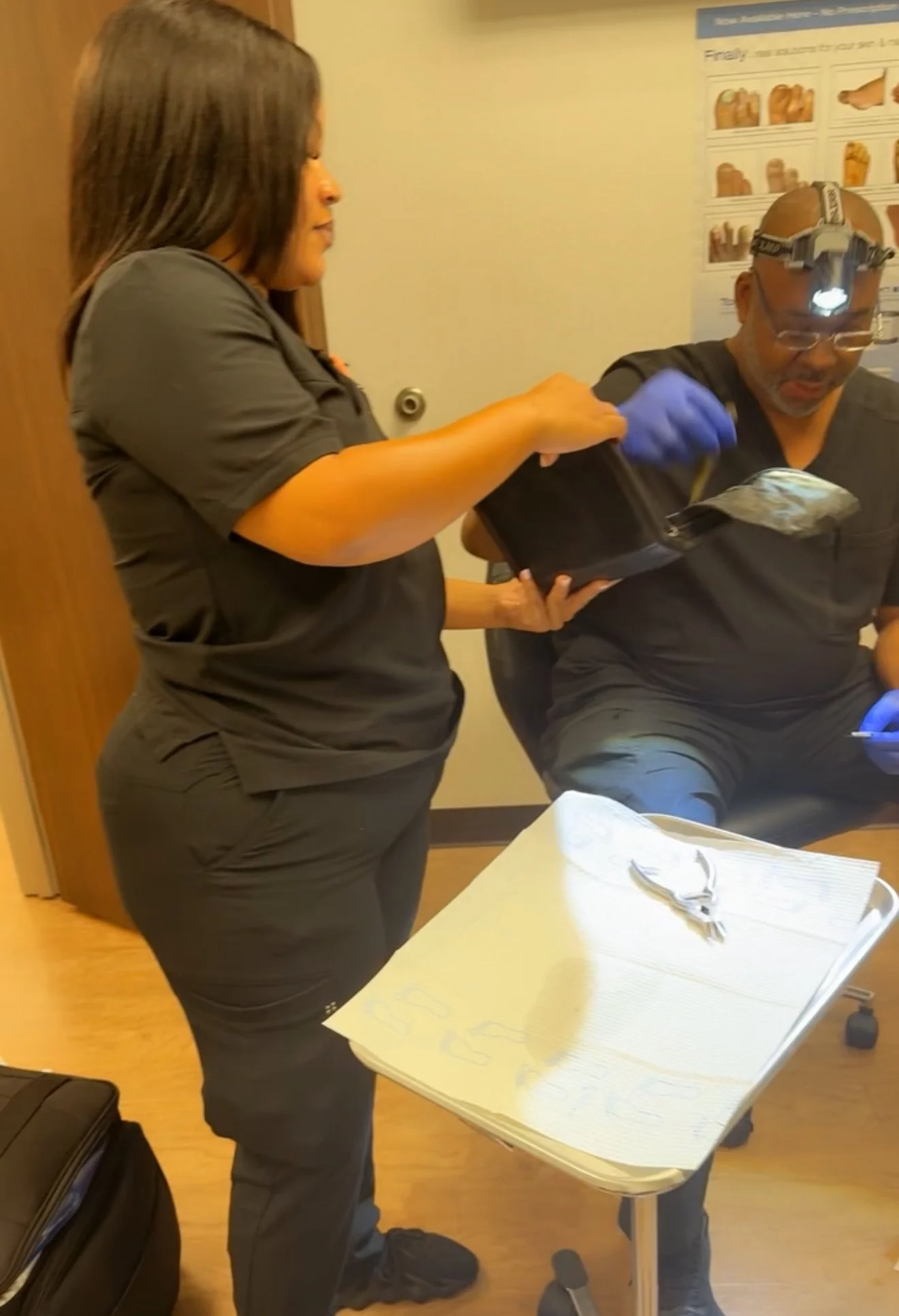 Two healthcare professionals, a woman and a man, are in a room with a table and medical supplies. The woman is handing a small package or sample to the seated man, who is wearing protective glasses and a headlamp. Both are dressed in dark scrubs, and there is a poster with illustrations on the wall behind them.