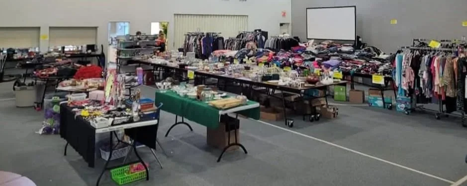 Tables and racks filled with various secondhand clothing, accessories, and small household items at a flea market or thrift sale in a large indoor space.
