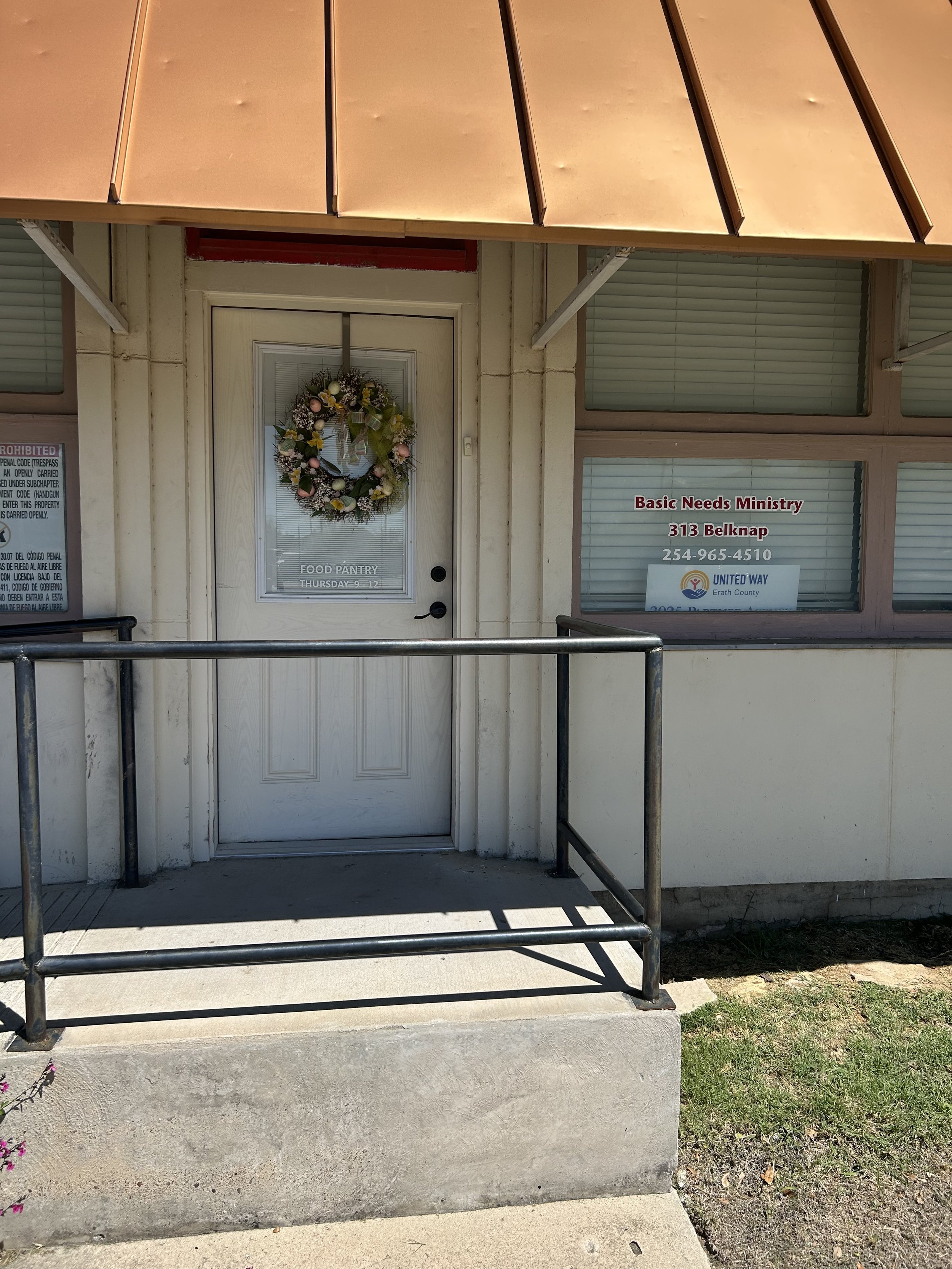 A storefront with a white door decorated with a floral wreath, displaying a sign for a food pantry. To the right, there's a window with signs for Basic Needs Ministry and United Way, with contact information. A small ramp with metal railings leads to the door under a tan awning.