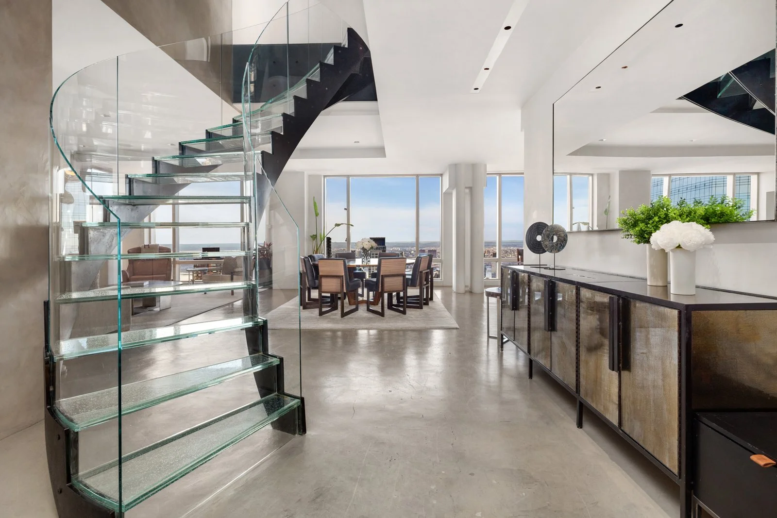 Modern apartment interior with glass staircase, a dining area with a round table and chairs, a sideboard with decorative items, and large windows offering a city view.