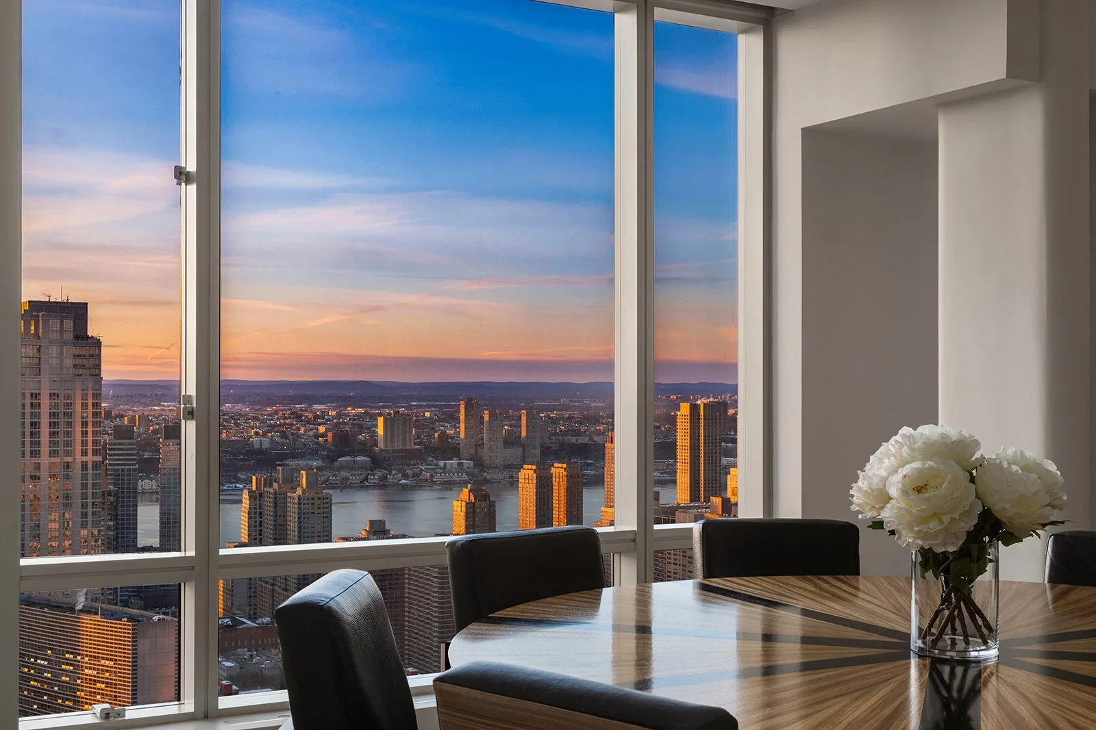 City skyline view of tall buildings and a river at sunset from a modern dining room with a round wooden table, black chairs, and a vase of white flowers.