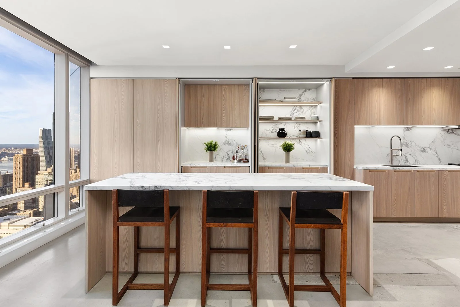 Modern kitchen with white marble countertops, light wood cabinetry, three black chairs at a marble island, large window with city view, open shelves with plants and dishes, sink with a faucet.