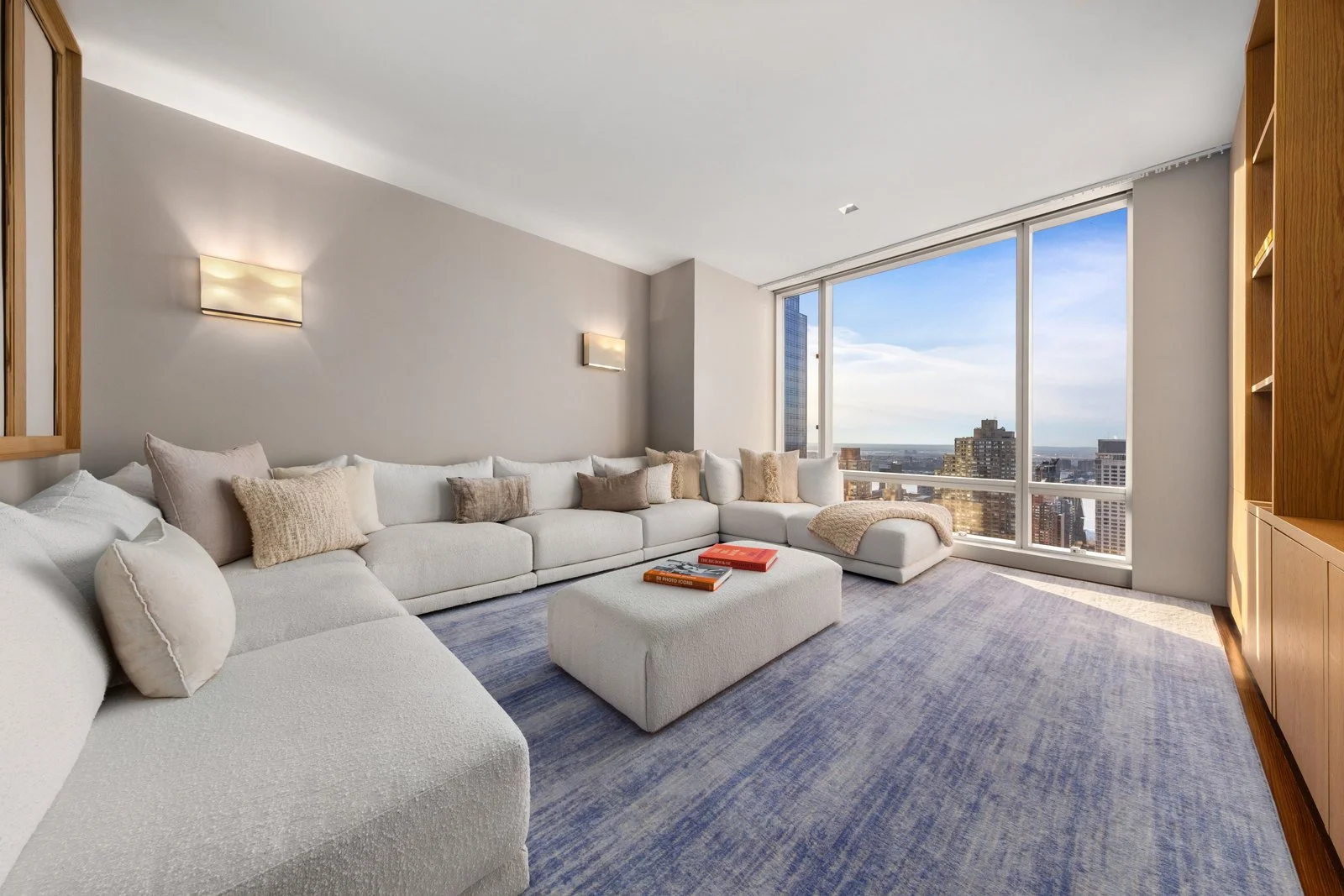 Modern living room with large white sectional sofa, beige and brown throw pillows, a white ottoman with books, a blue area rug, and floor-to-ceiling windows showcasing a city skyline with tall buildings and a bright sky.