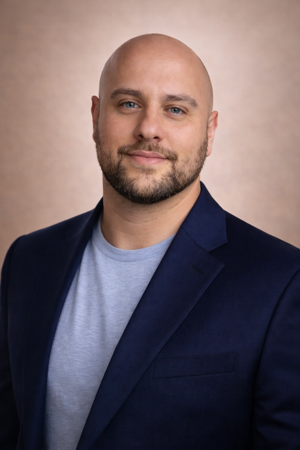 Headshot of a bald man with a trimmed beard, wearing a navy blazer and light gray t-shirt, smiling softly against a neutral background.