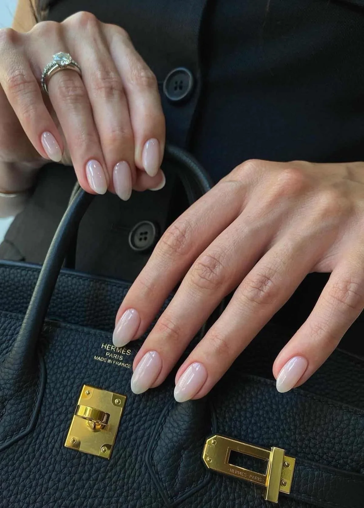 Mains féminines posant sur un sac à main Hermès noir avec fermoirs en or, on porte une bague en diamant et des ongles ovales en vernis clair.