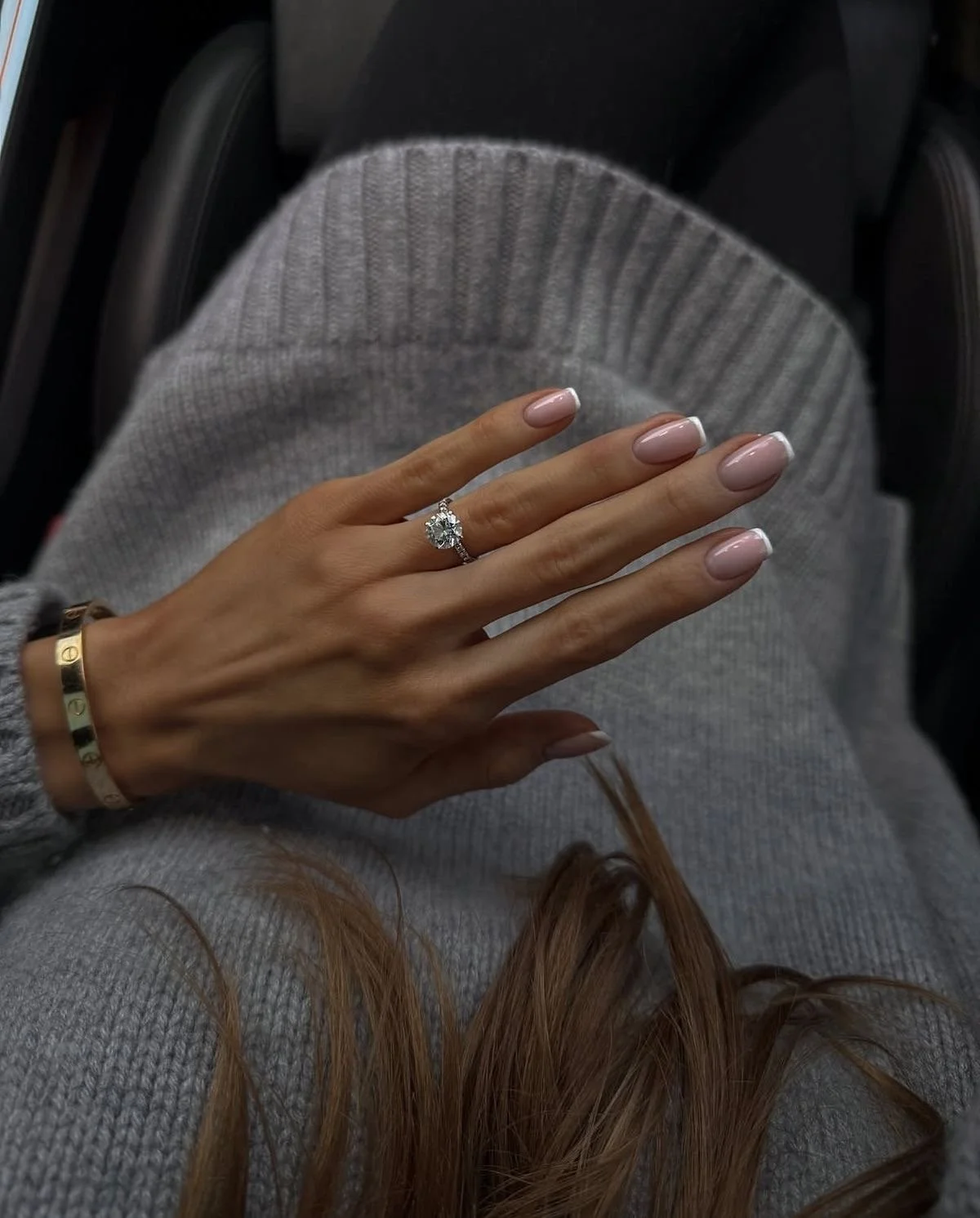 Main avec bague de fiançailles portant une pierre précieuse. La main repose sur un vêtement en maille gris.
