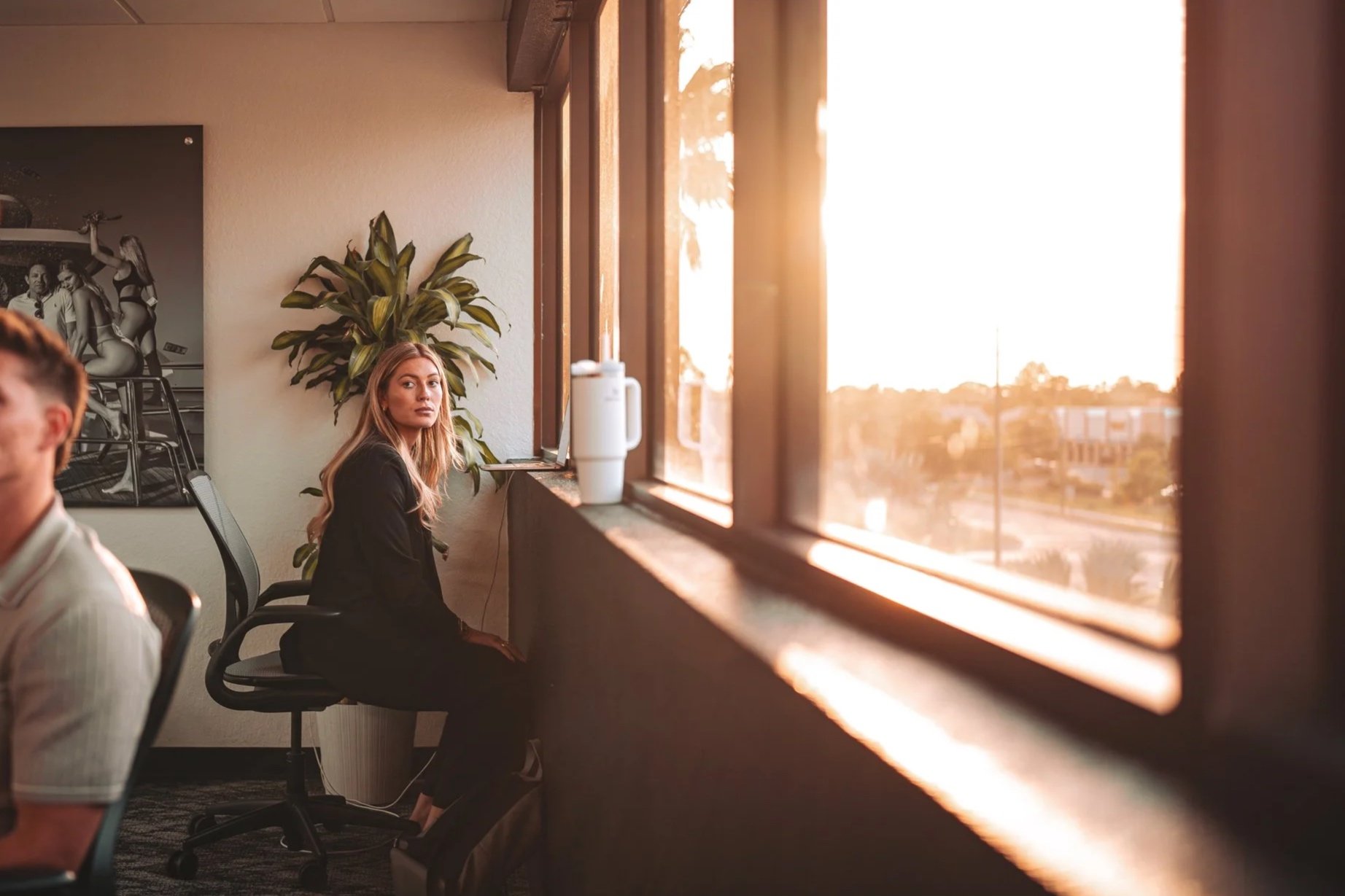 A young woman sitting on an office chair by a large window during sunset, with a potted plant behind her and a framed black-and-white picture on the wall.