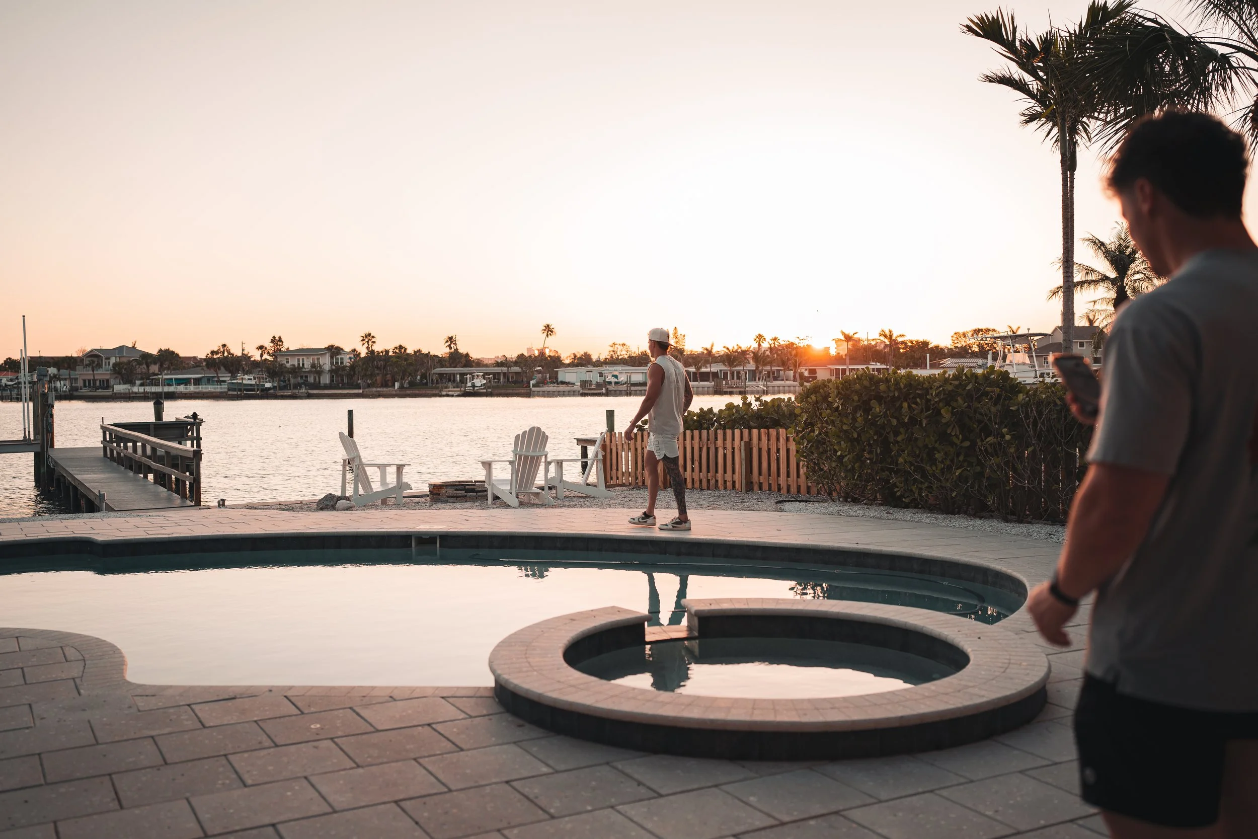 Two young men near a pool at sunset, one is using a phone while the other is walking along the waterfront with palm trees and houses in the background.