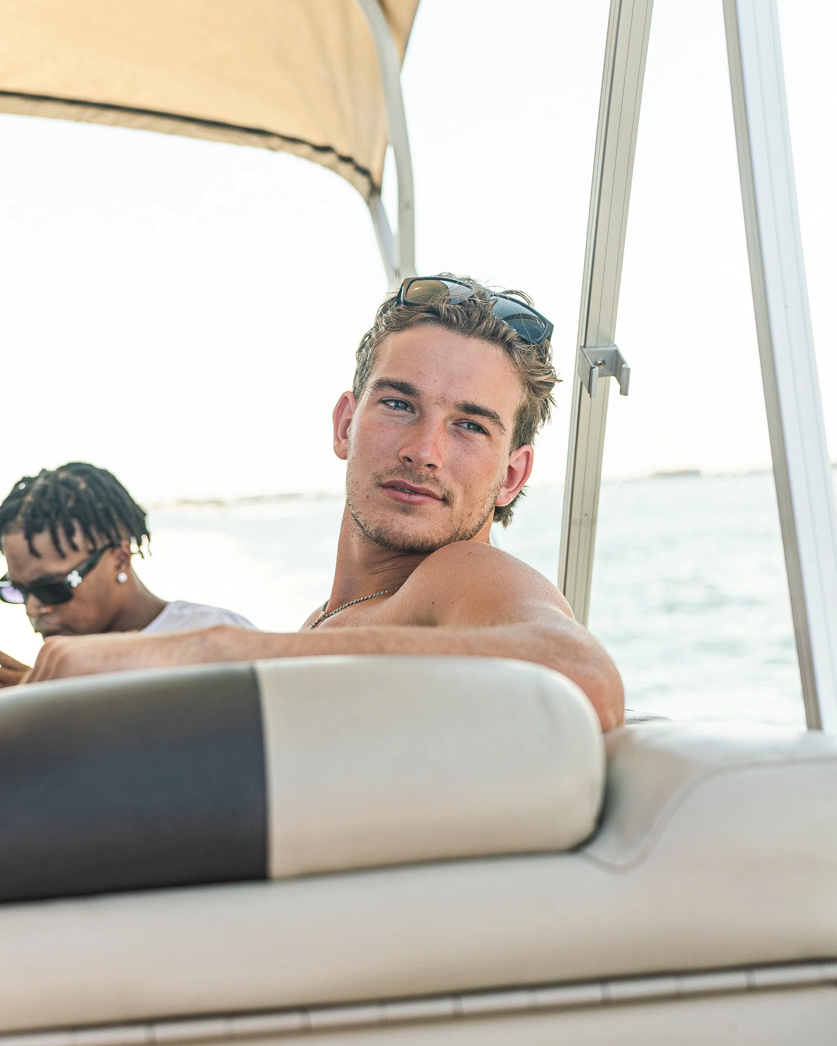 A young man with sunglasses on his head relaxing on a boat with water in the background, and another person with dreadlocks and sunglasses in the background.