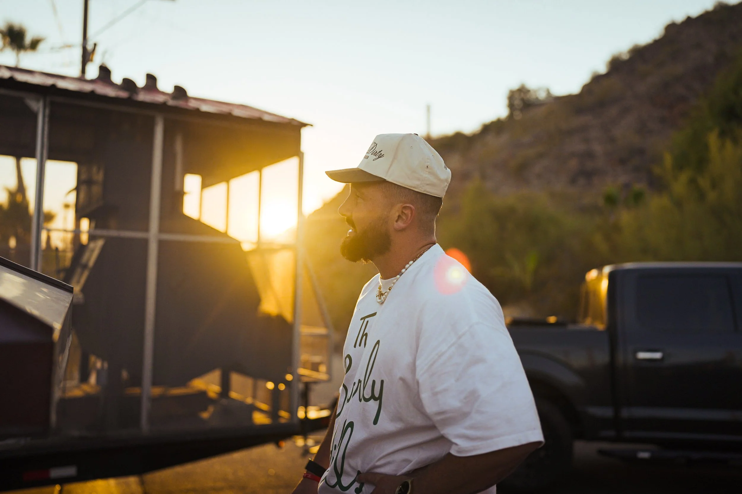 A man with a beard and baseball cap smiling and standing outdoors during sunset, with a black truck and hillside in the background.