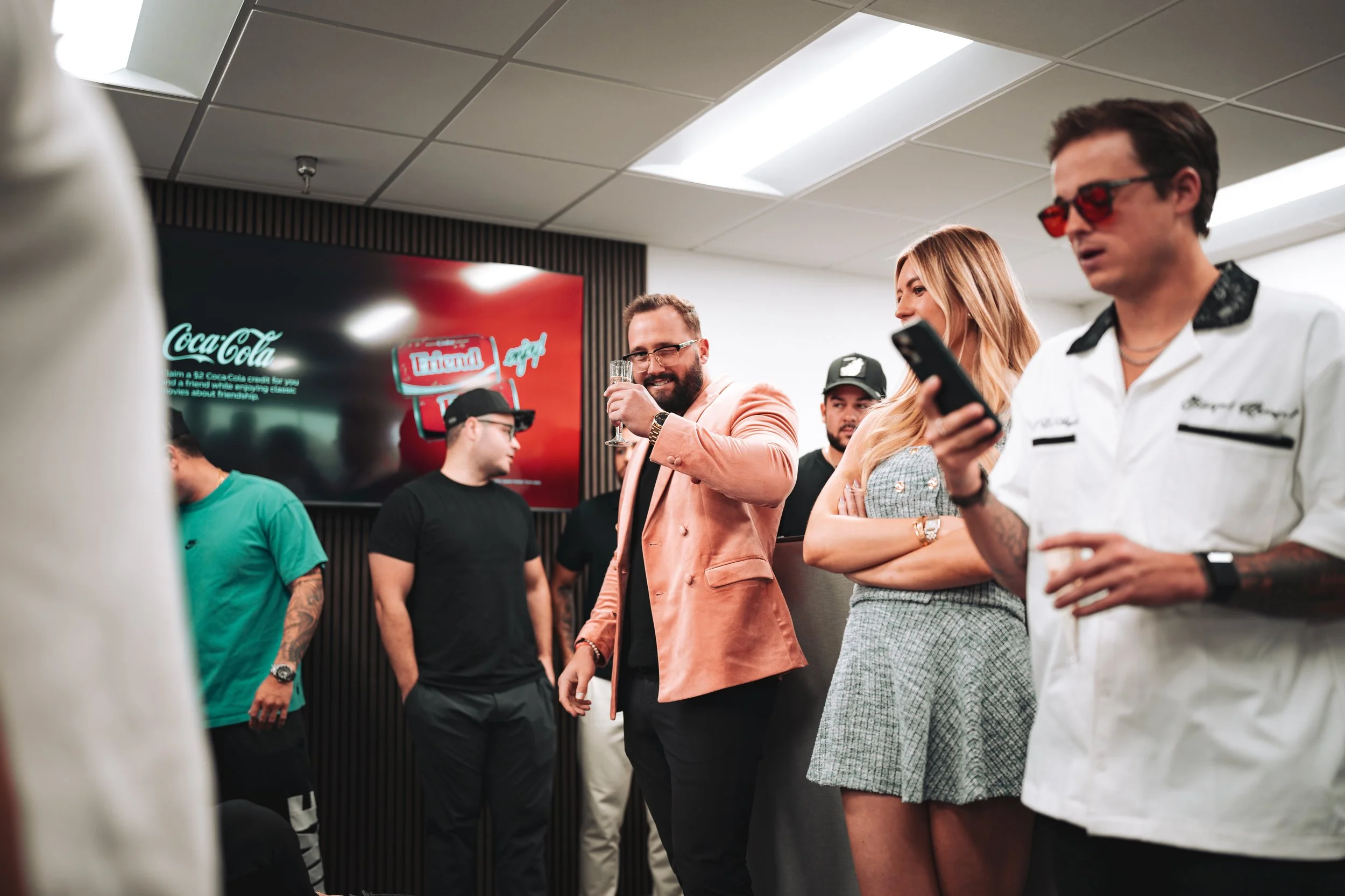 Group of people at a social gathering, some people are looking at their phones, one man with sunglasses holding a drink, a woman is smiling and looking at her phone, a man in a pink blazer is smiling and holding a glass, and others are standing aroun