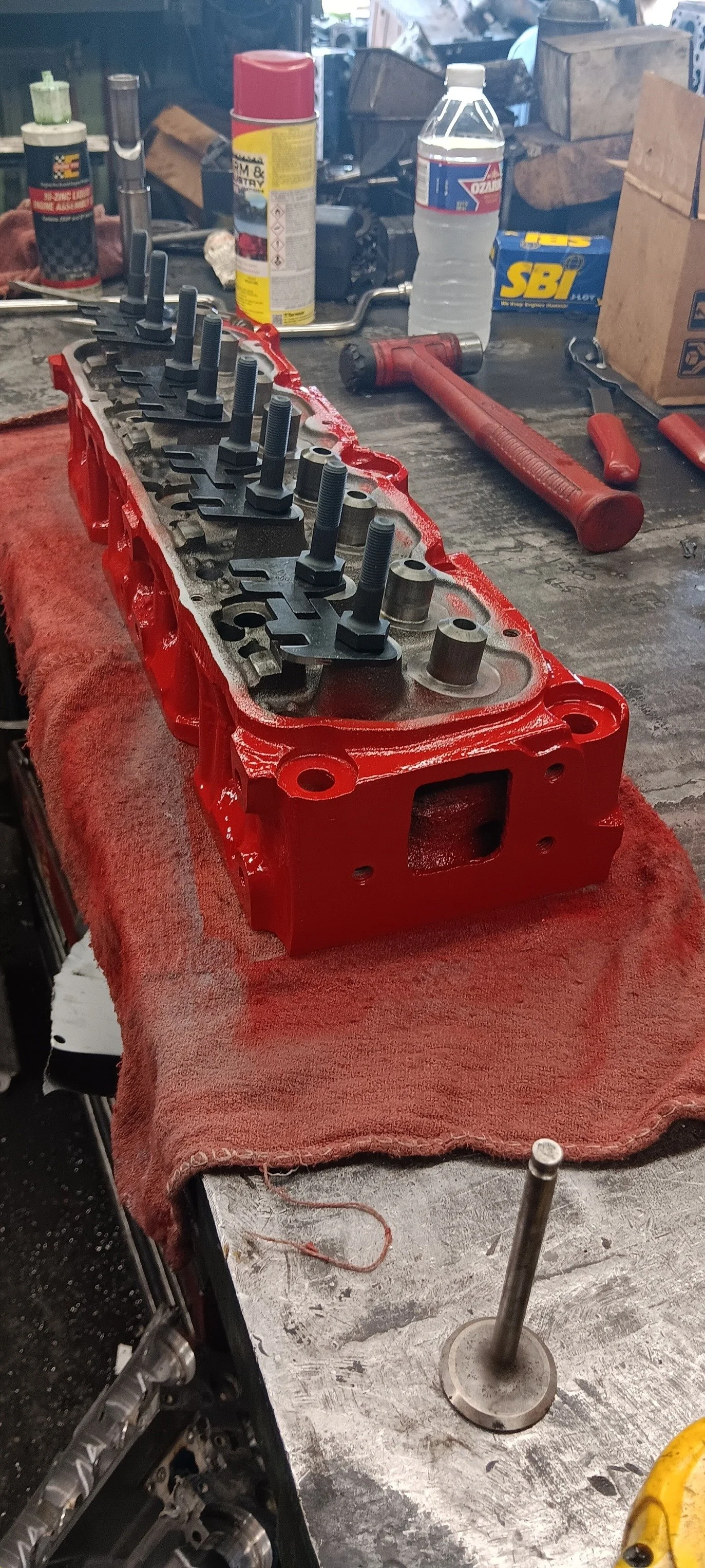 An engine cylinder head on a workbench with tools and supplies in the background.