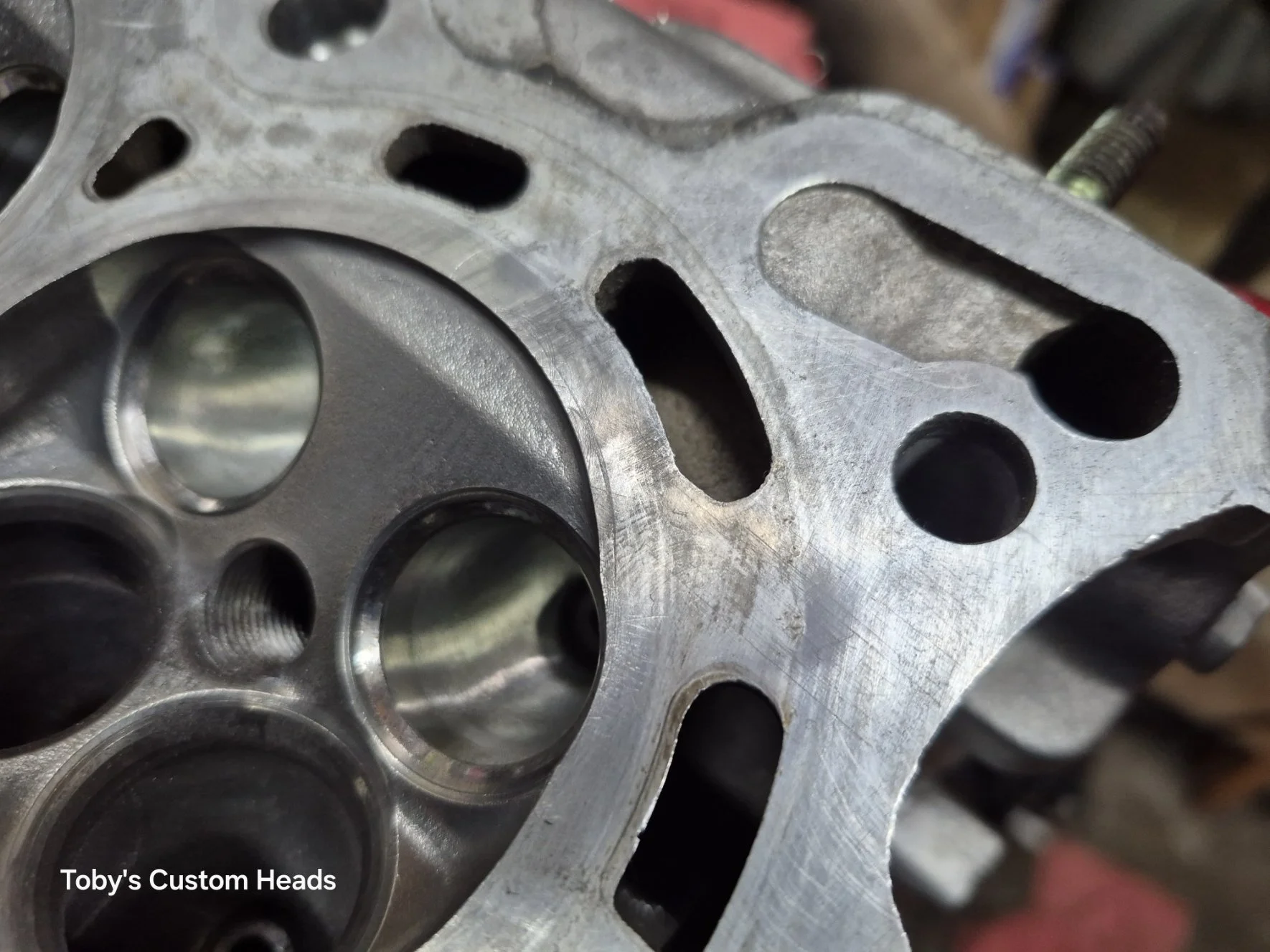 Close-up of a metal engine cylinder head with machined surfaces and valve seats, labeled 'Toby's Custom Heads'.
