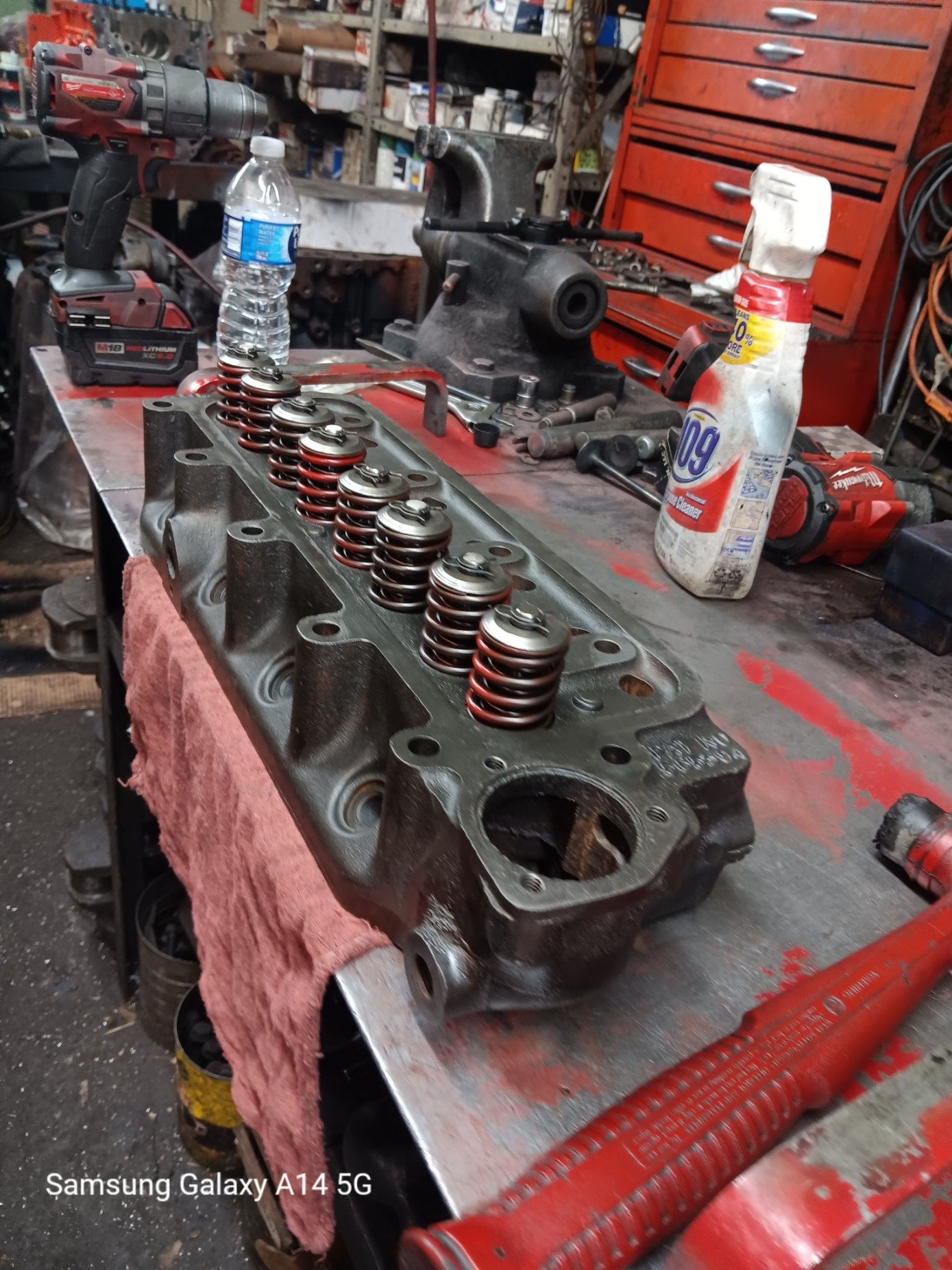 A disassembled engine cylinder head with valve springs on a workbench in a garage workshop.