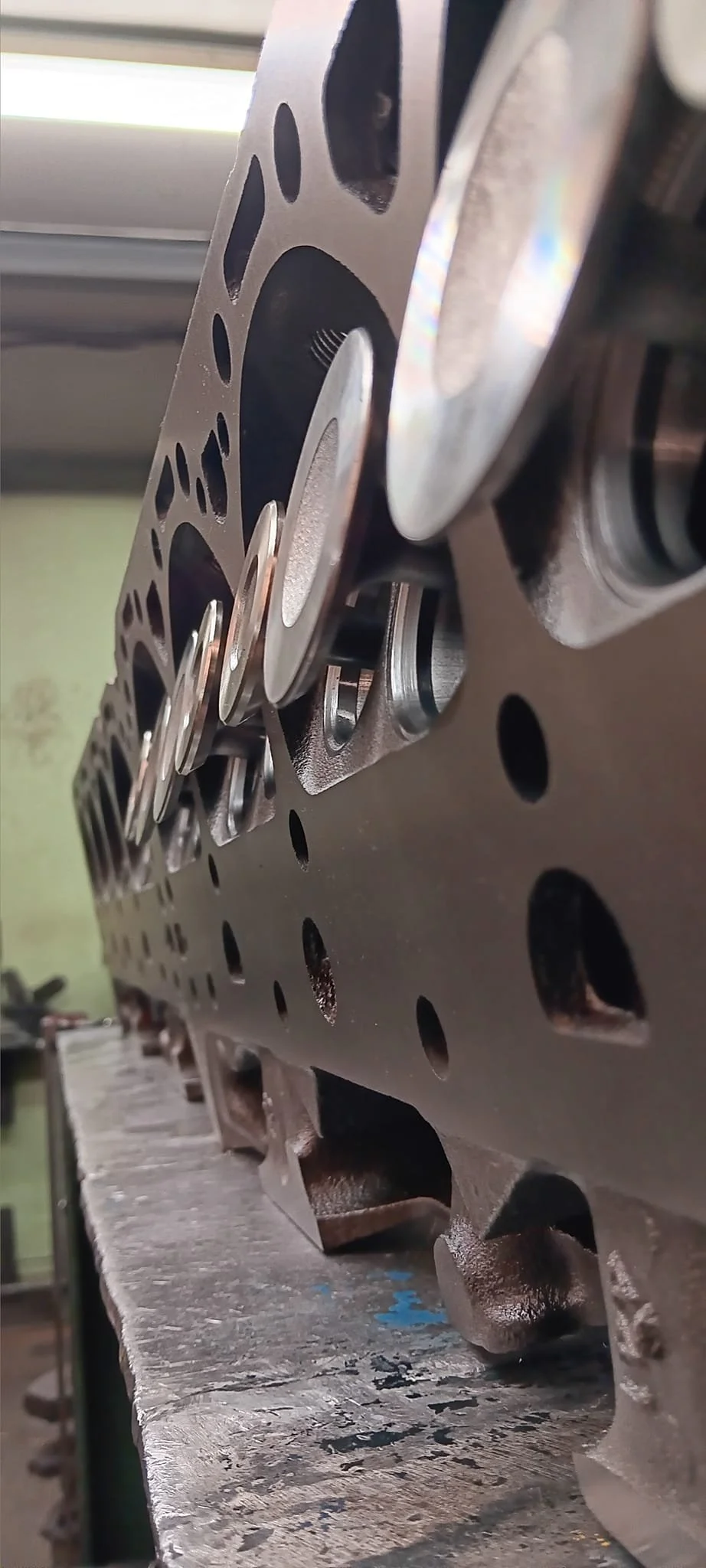 Close-up view of a metal engine cylinder head with valves and springs, resting on a workbench in a workshop.