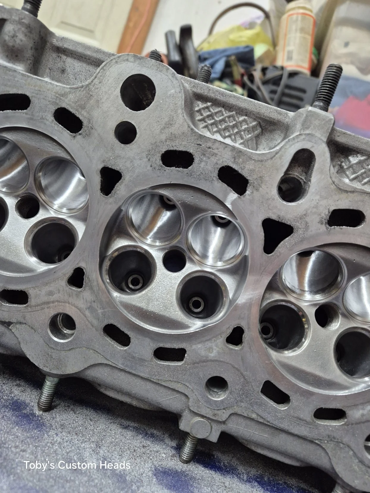 Close-up of a silver engine cylinder head with machined ports, bolts, and cutouts, placed on a workbench in a garage or workshop.
