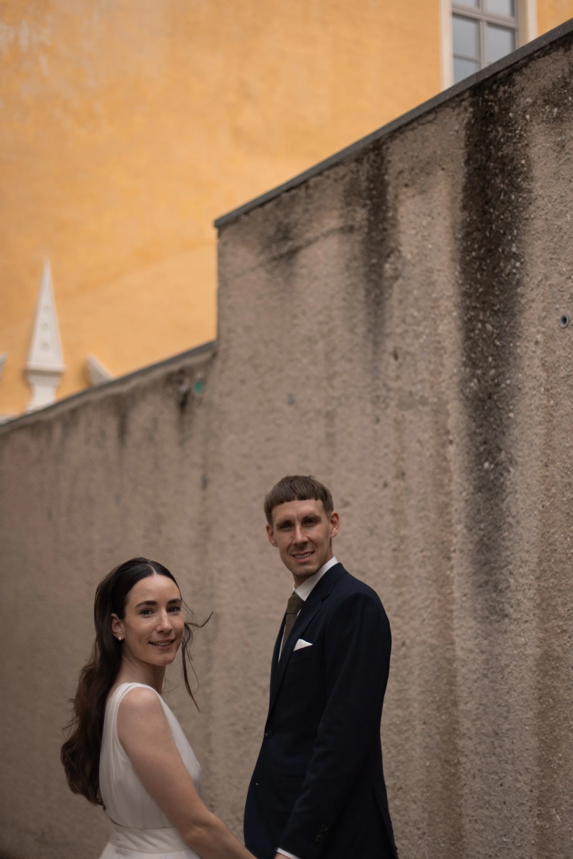 A young couple dressed in wedding attire walking outdoors, smiling and holding hands, with a yellow building and a window in the background.