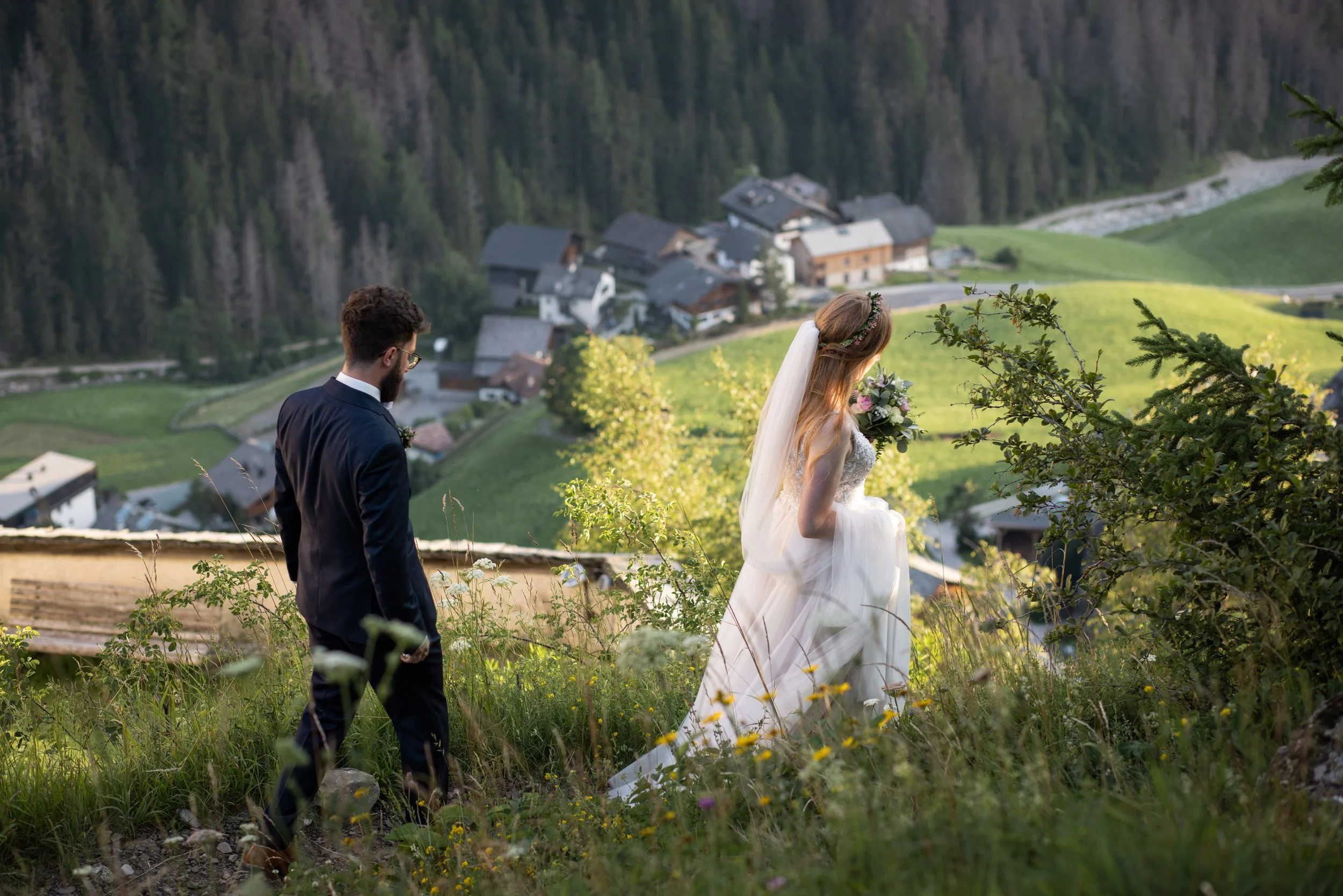 A bride in a wedding dress and floral headpiece and a groom in a suit walking on a path. Near La Val in Val Badia, with the Dolomites and forest in the background.