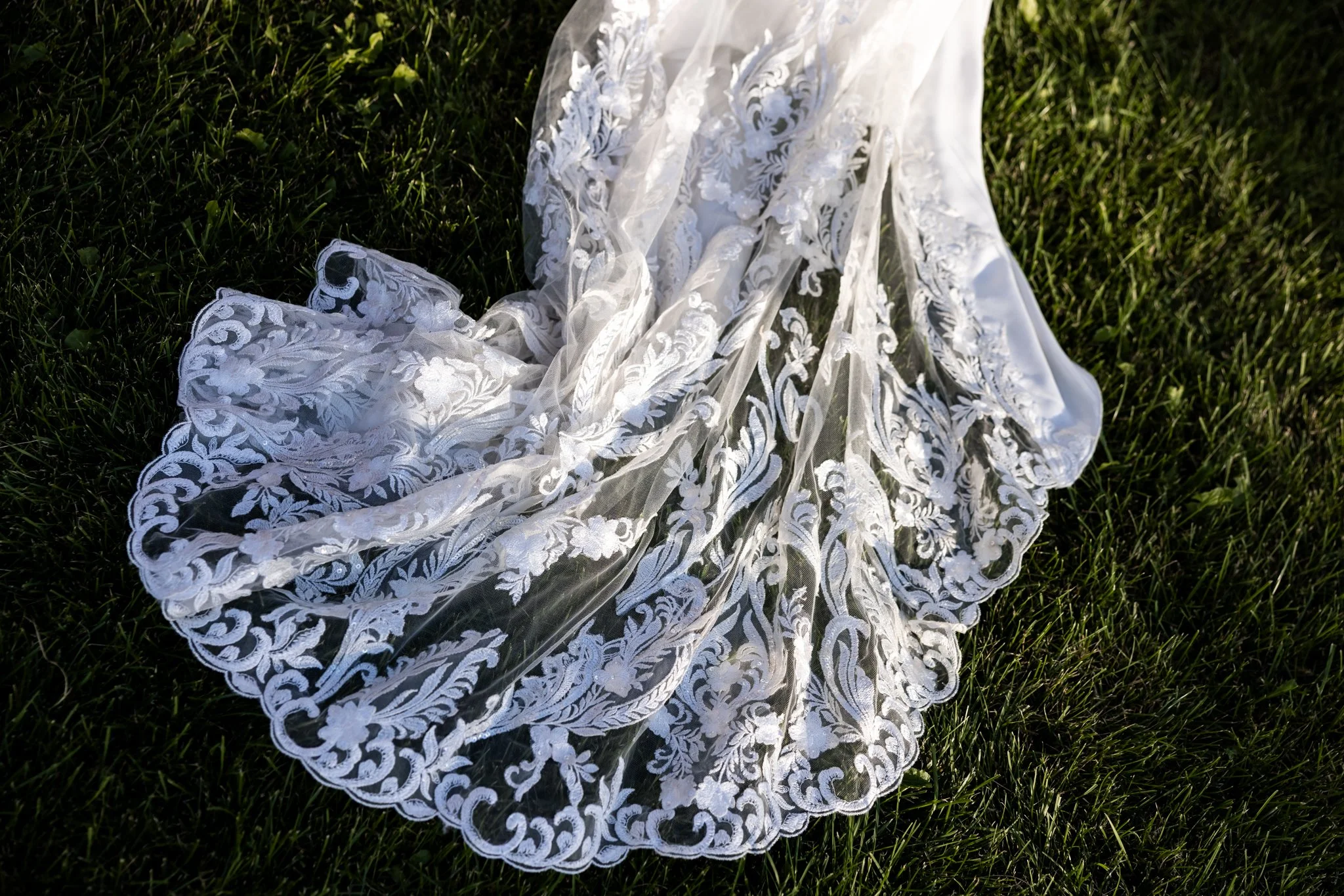 A white lace wedding veil with intricate floral embroidery laid out on green grass.