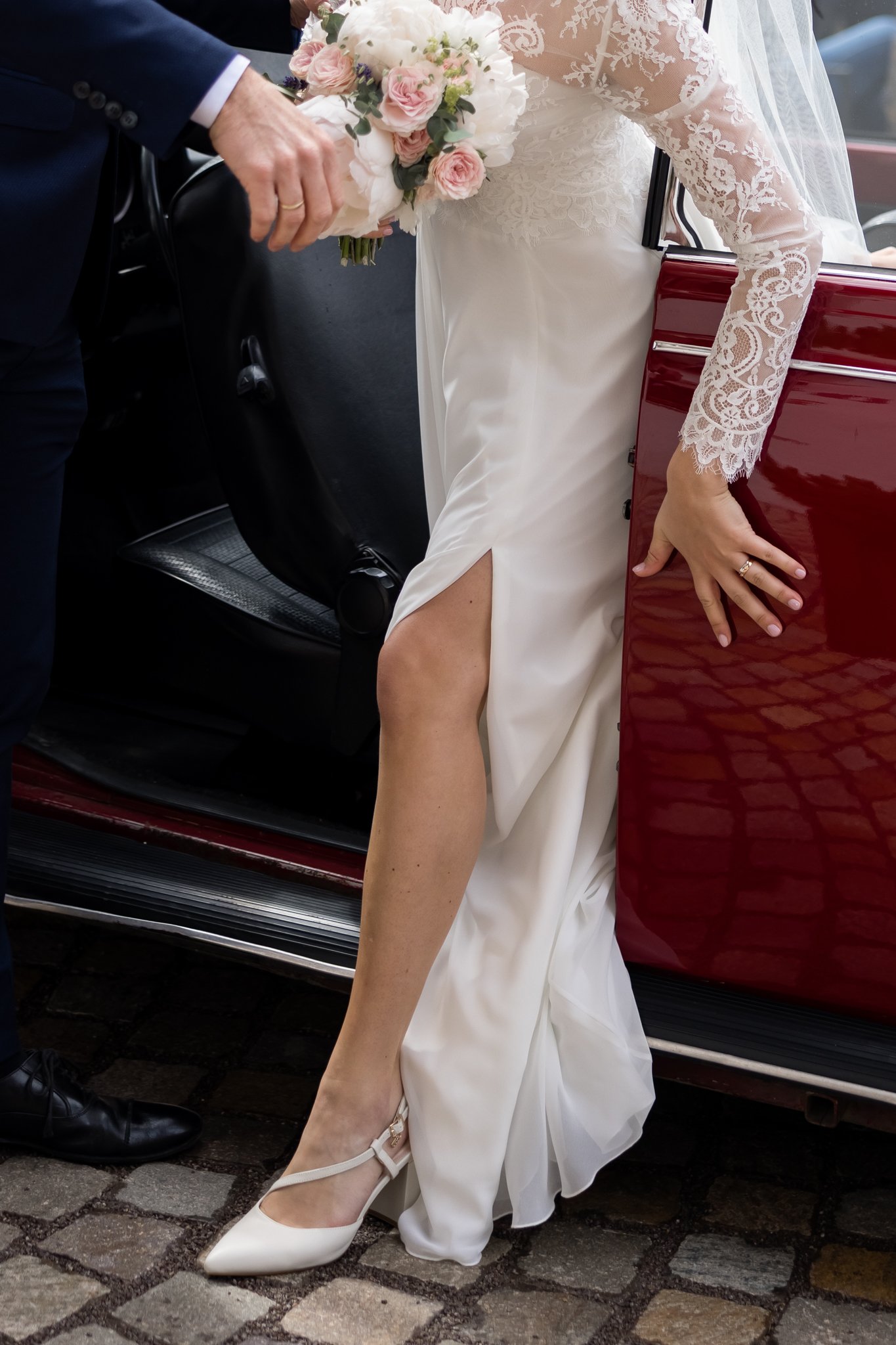 A bride wearing a lace wedding dress and white heels is seated in a vehicle, holding a bouquet of pink and white flowers, with a groom in a navy suit assisting her, all on cobblestone pavement.