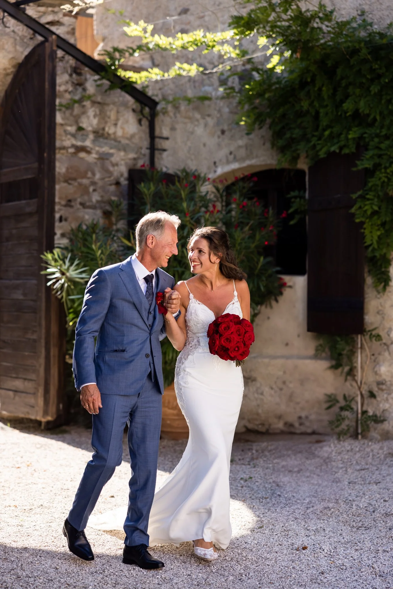 A bride in a white wedding gown holding a bouquet of red roses, walking with the groom in a blue suit, into a magnificent wedding location Castel Fahlburg in South Tyrol.