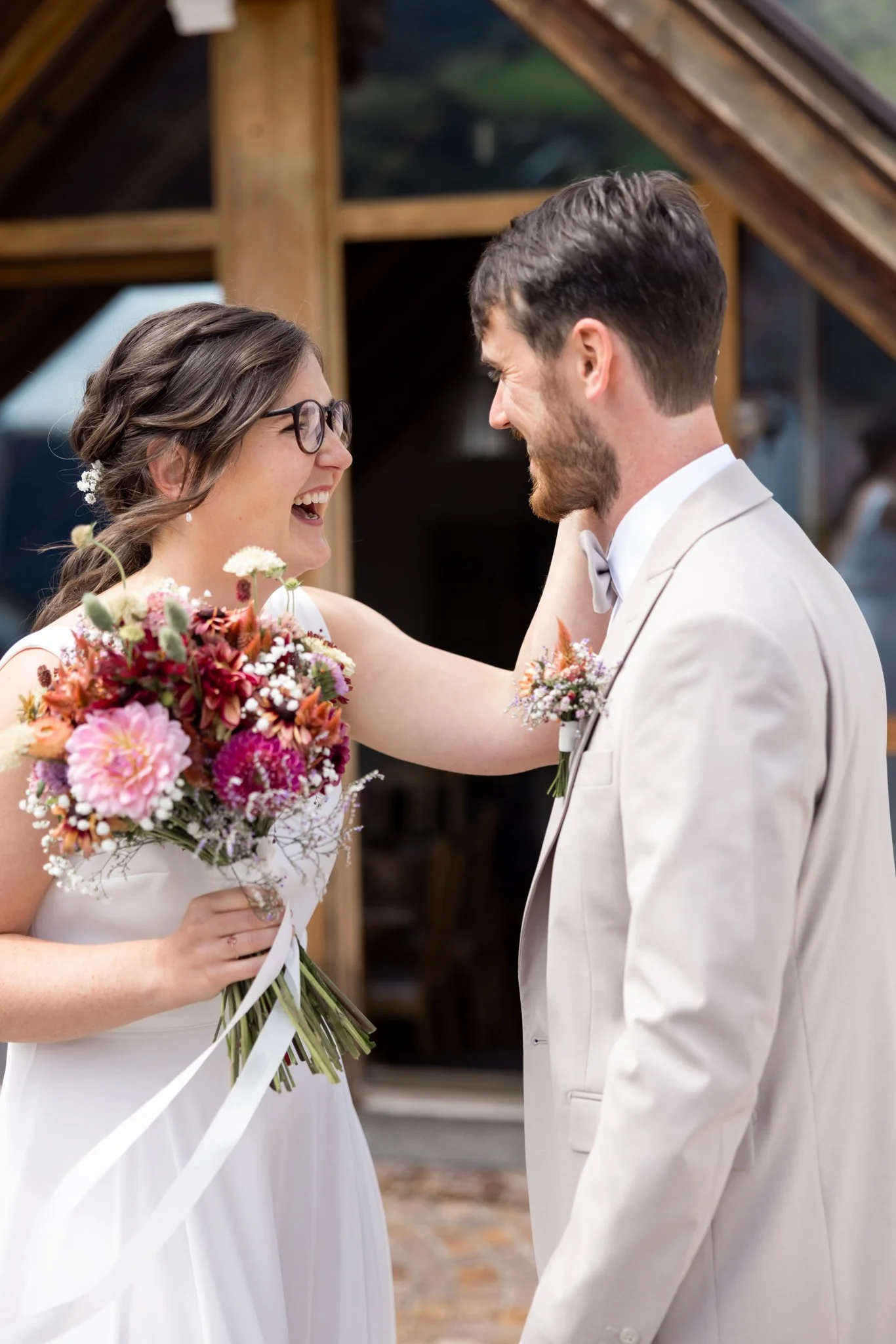 A bride and groom sharing a joyful moment at their wedding, with the bride holding a bouquet of flowers and wearing glasses, and the groom dressed in a light-colored suit, both smiling and looking at each other.