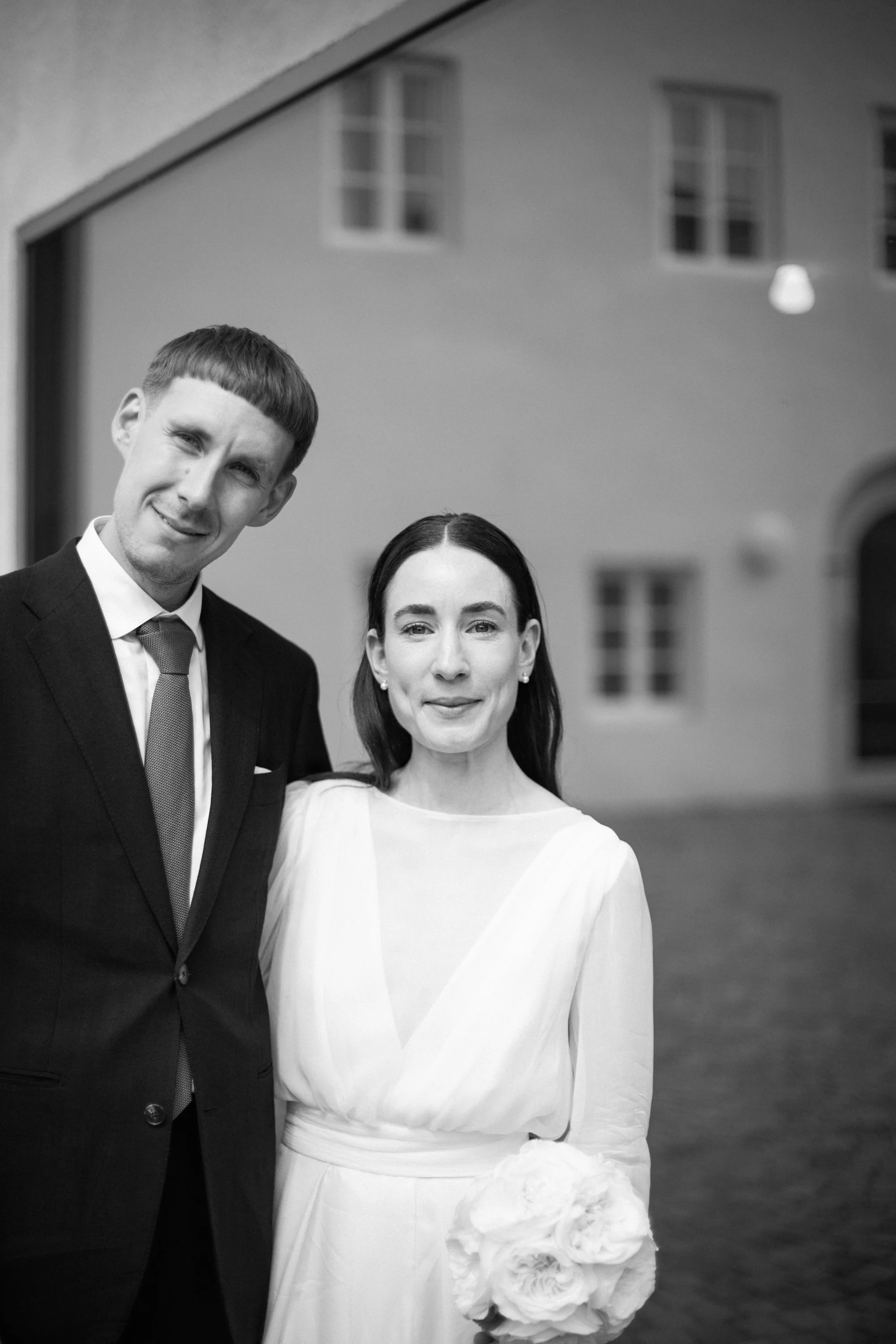 Black and white photo of a man in a suit and a woman in a dress standing close together, smiling, indoors with windows and a ceiling light in the background.