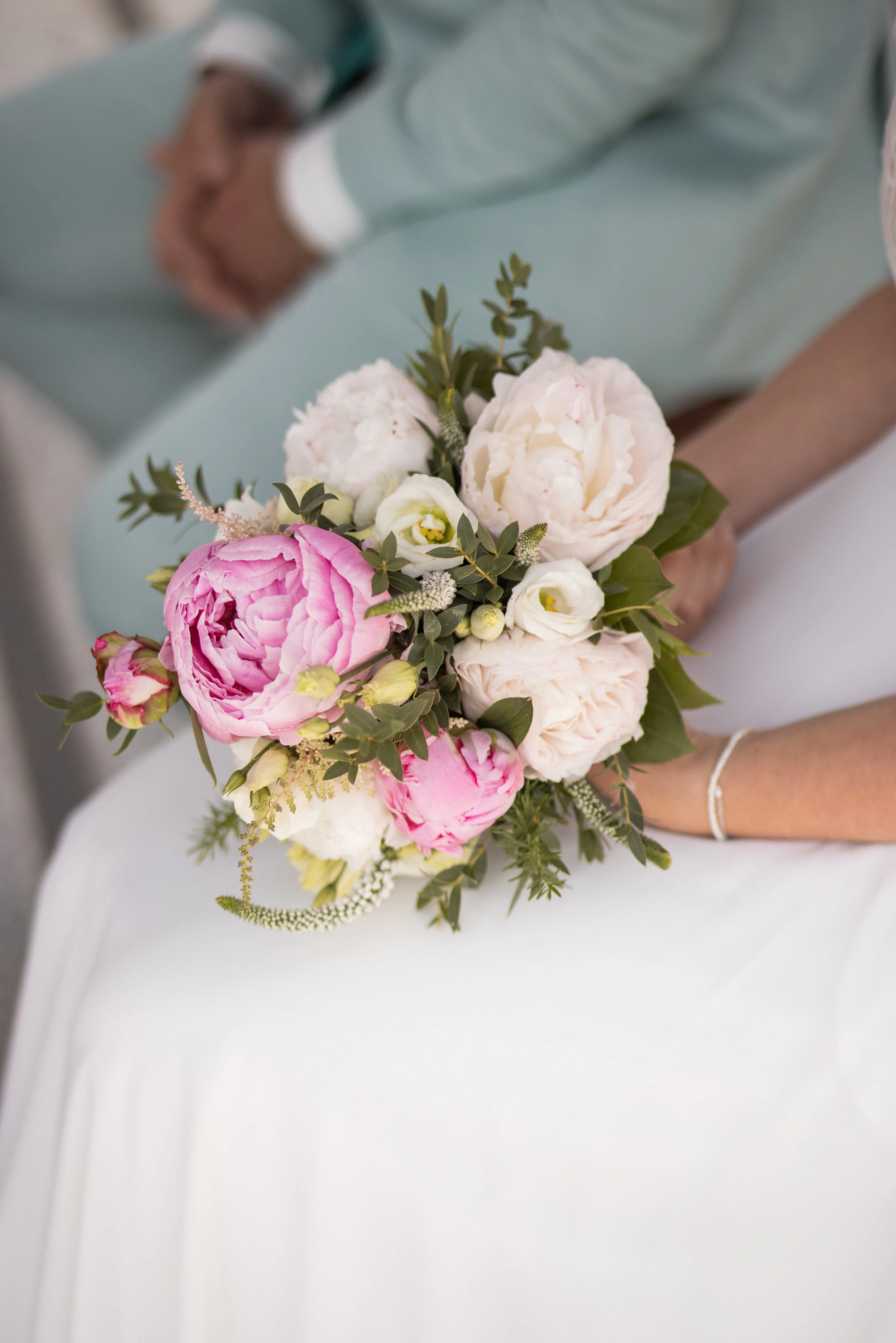Close-up of a bride's bouquet featuring pink and white peonies, white lisianthus, and greenery held by a bride in a white wedding gown.